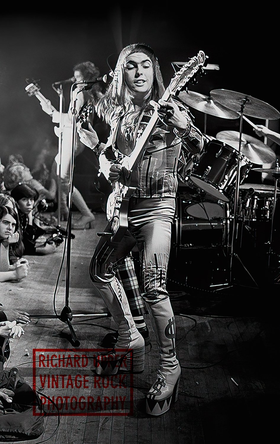 Black and white photograph of a woman playing an electric guitar on stage, with a drum set in the background. Audience members are seated on the floor in front of her, watching the performance.