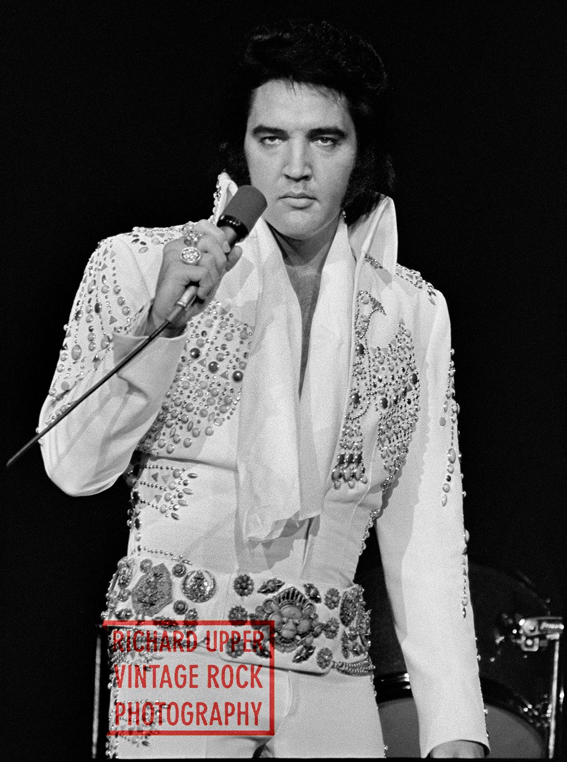 Black and white photograph of Elvis Presley holding a microphone, wearing a white jumpsuit adorned with studs and embellishments, on stage.