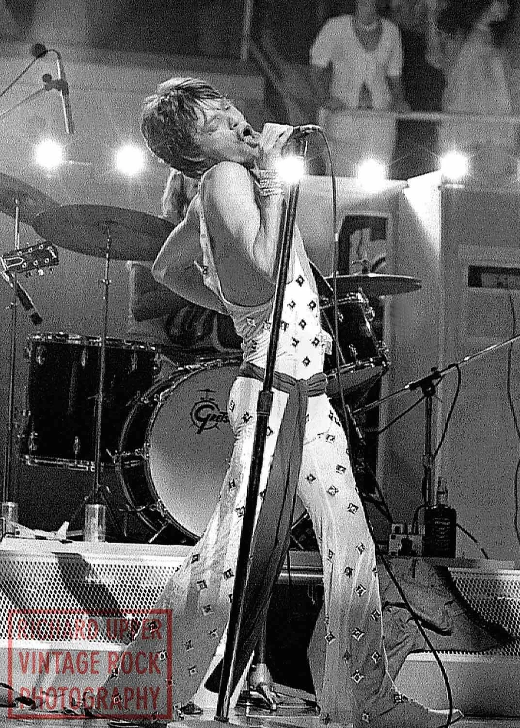 A female singer passionately performing on stage with a microphone, wearing patterned pants and a sleeveless top, in front of a drum set. There are a few people in the background watching.