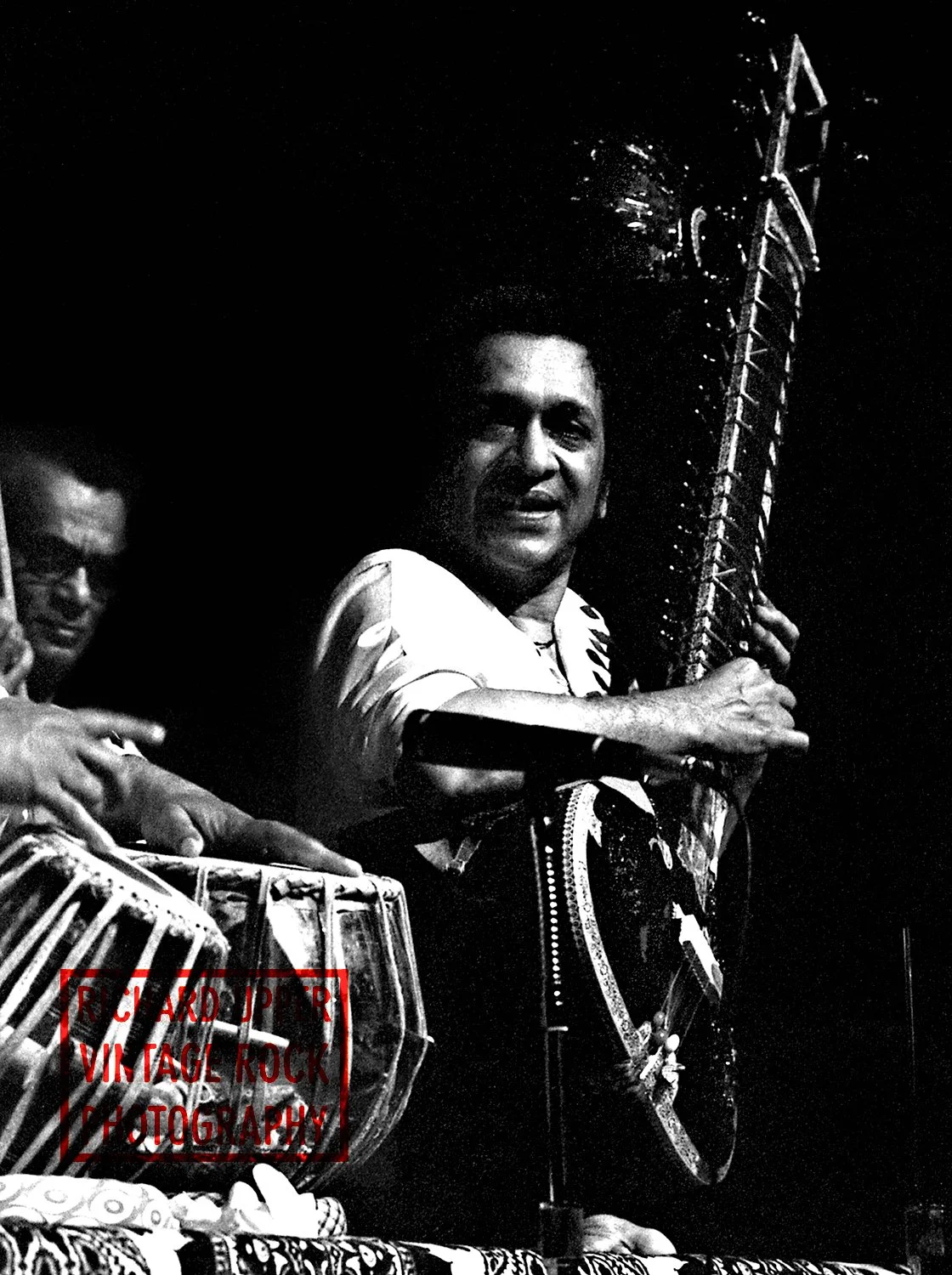 A man playing a drum and another man holding a traditional stringed instrument on stage at night.