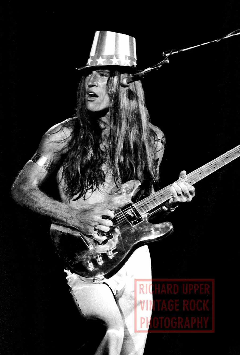 A man with long hair playing an electric guitar, wearing a tall hat decorated with stars, during a performance, in black and white.