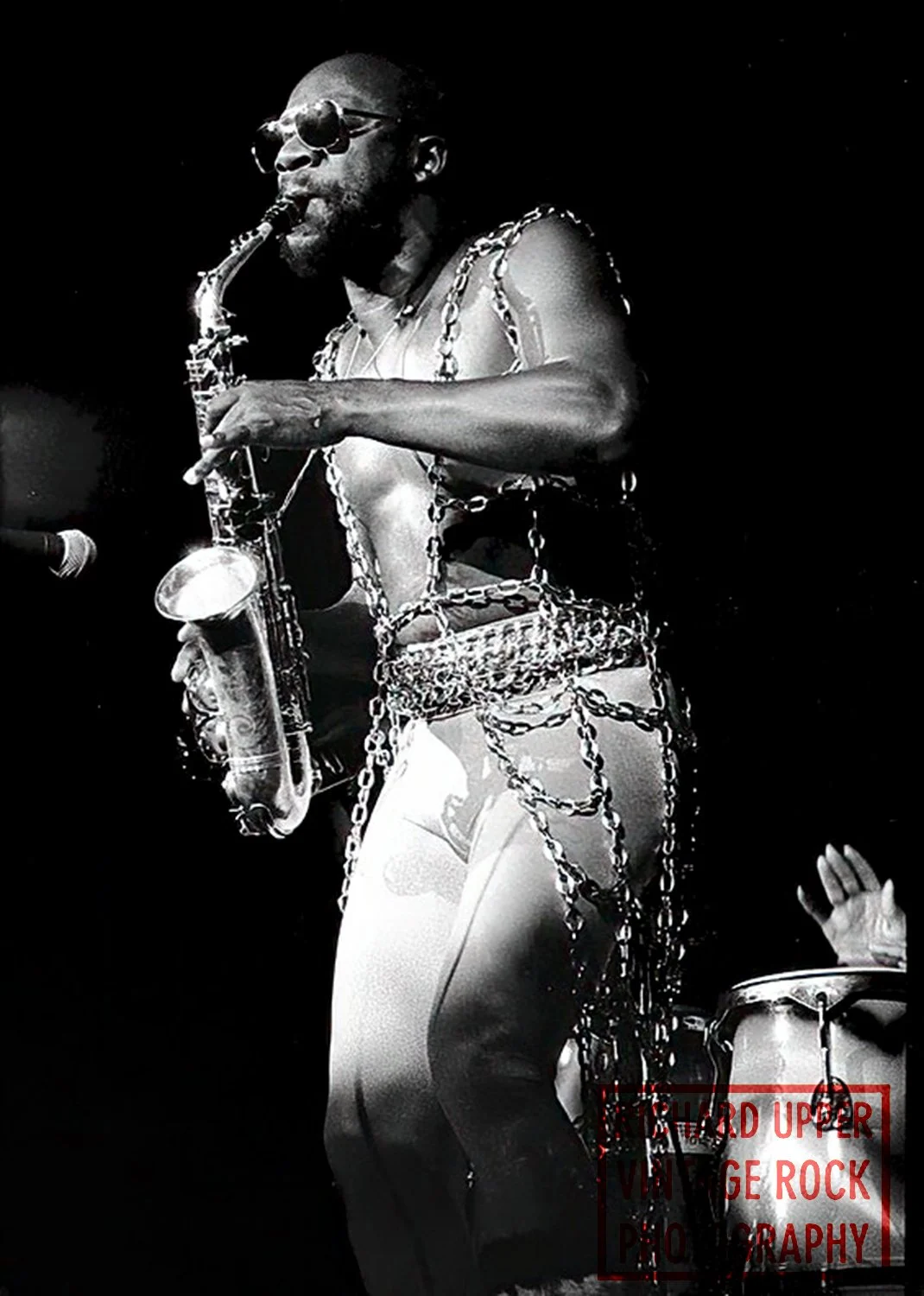 A black and white photo of a man playing a saxophone on stage. The man is shirtless, wearing sunglasses, and adorned with numerous chains and accessories. There is a drum set part visible in the lower right corner with a hand raised near it.