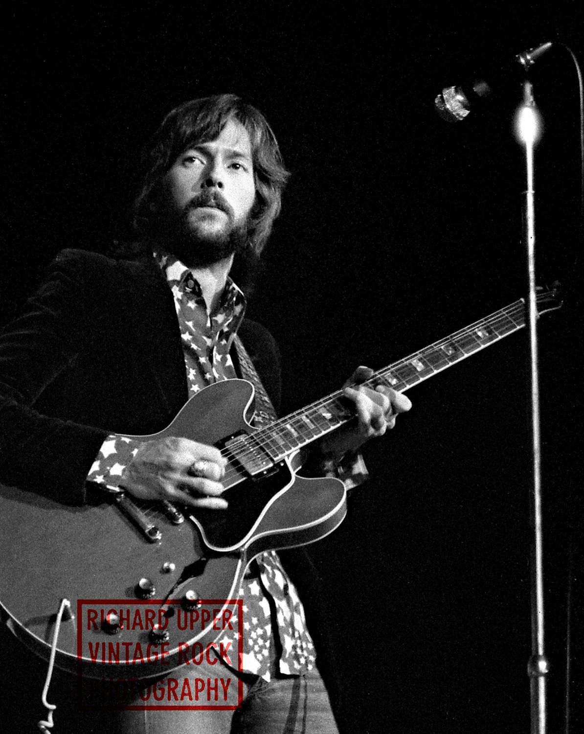 Black and white photo of a male musician playing an electric guitar on stage, with a microphone stand nearby and a dark background. The text 'Richard Upper Vintage Rock Photography' is overlaid on the bottom left corner.
