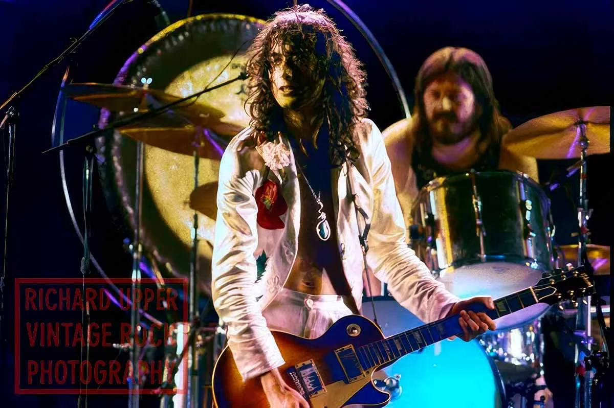 A musician playing an electric guitar and singing into a microphone on stage, with a drummer playing behind them, colorful stage lighting, and a large circular backdrop featuring an eye.