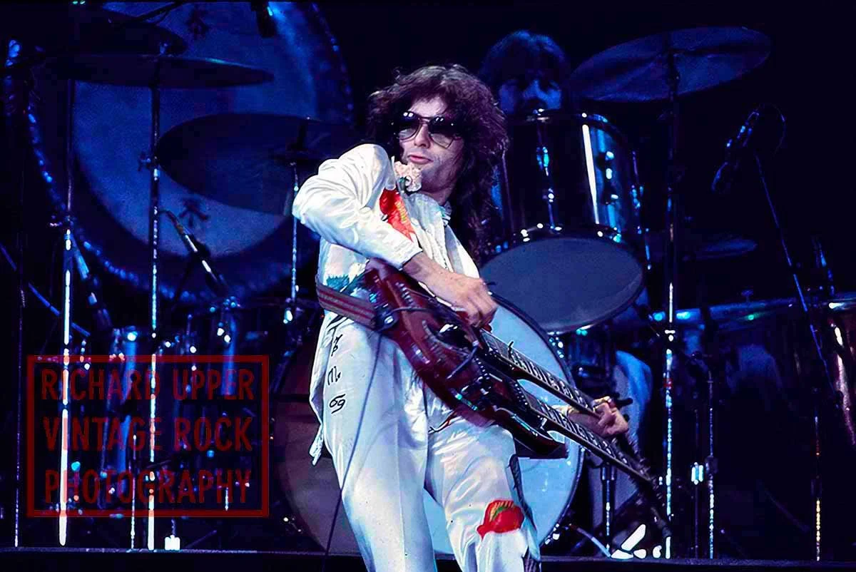 A male guitarist in white jumpsuit with colorful patches performs on stage with a drummer in the background. The guitarist has curly hair and wears sunglasses. The stage is dark with blue lighting, and the drummer is behind a large drum set.