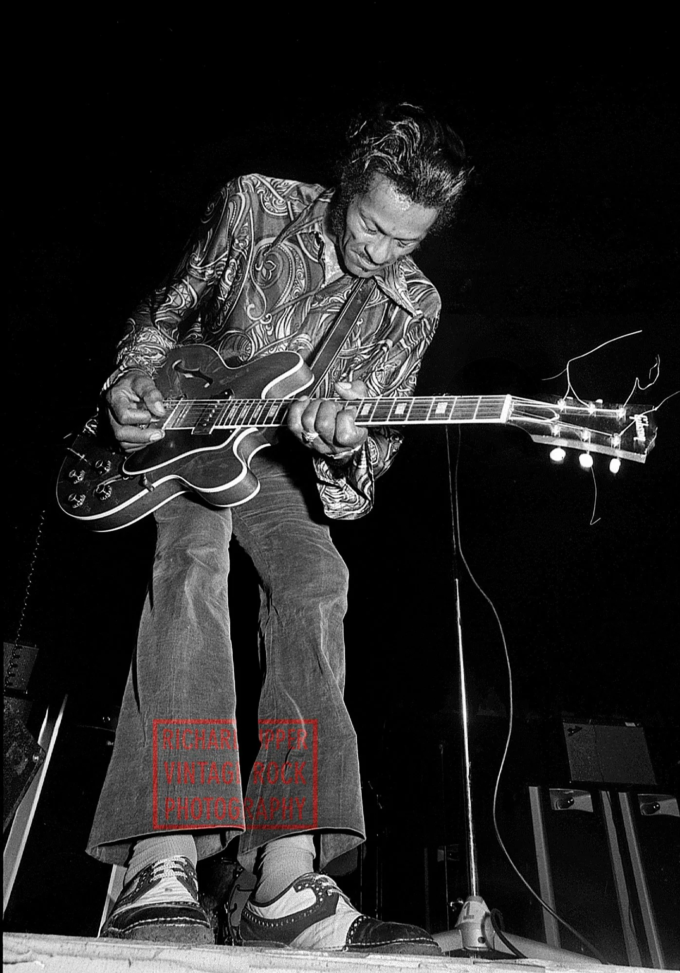 A man playing an electric guitar on stage, wearing a patterned shirt and jeans, with a dark background.