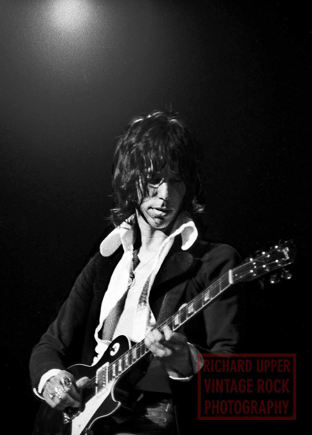 A black and white photograph of a musician playing an electric guitar. The musician has shaggy hair and is making a facial expression with their tongue out. There is a bright light above illuminating the scene and a red watermark in the lower right c