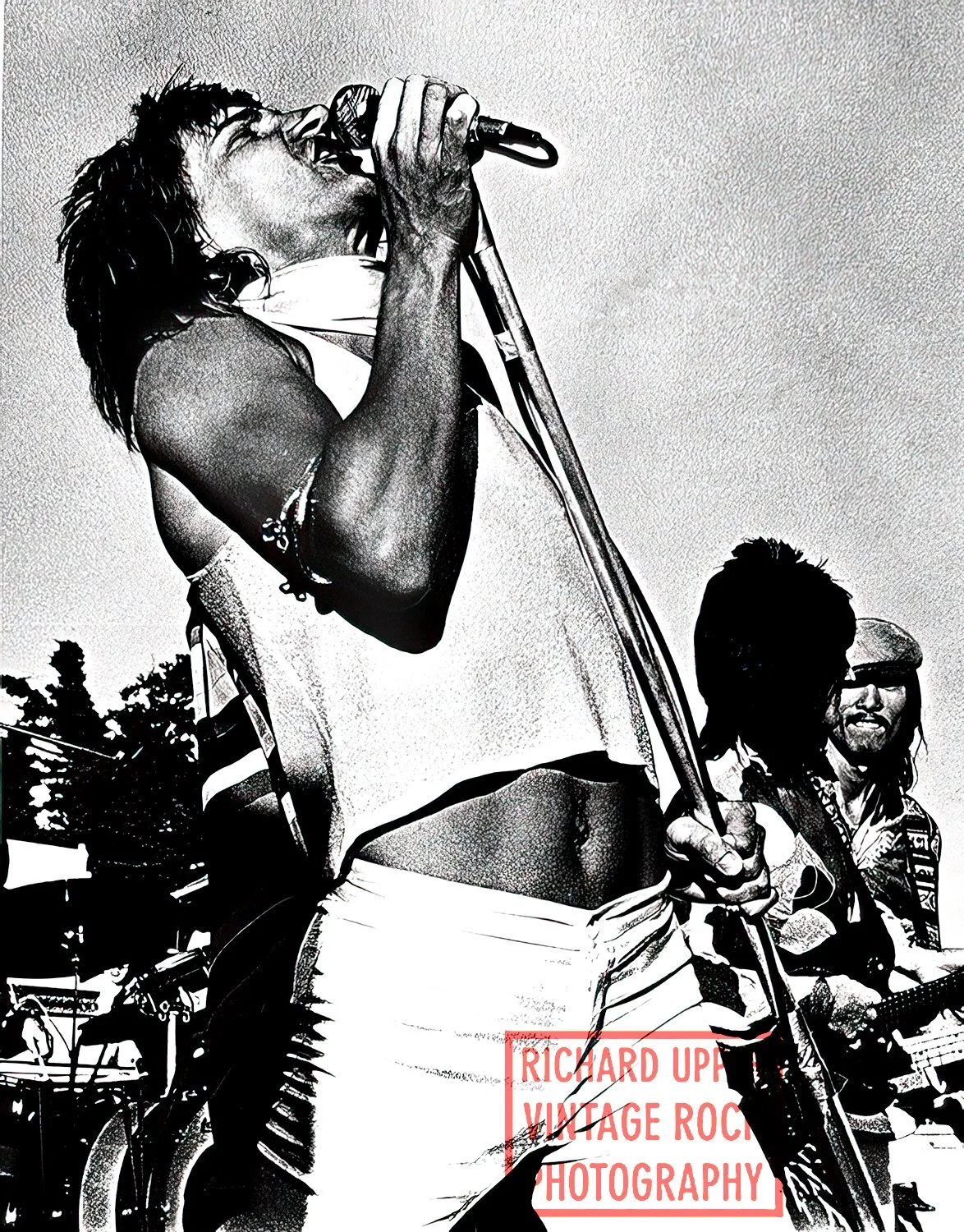 A black and white photograph of a man singing into a microphone at an outdoor event, with another person wearing sunglasses and a helmet in the background.