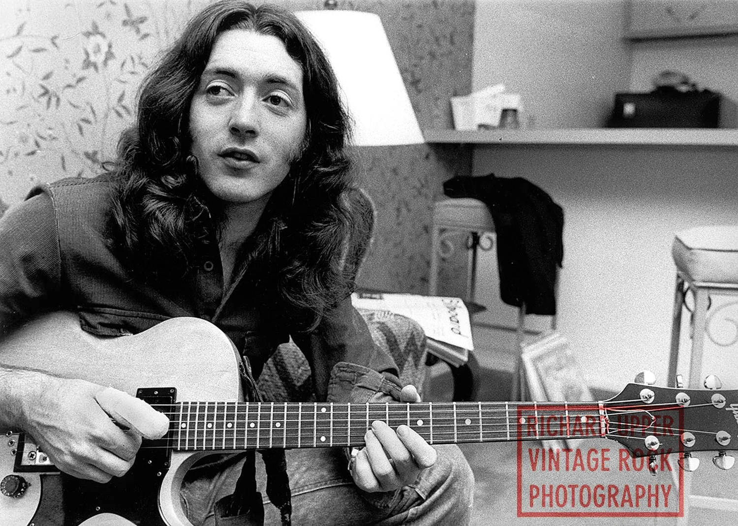 A woman with long, wavy hair, wearing a denim shirt, is sitting indoors and playing an acoustic guitar.