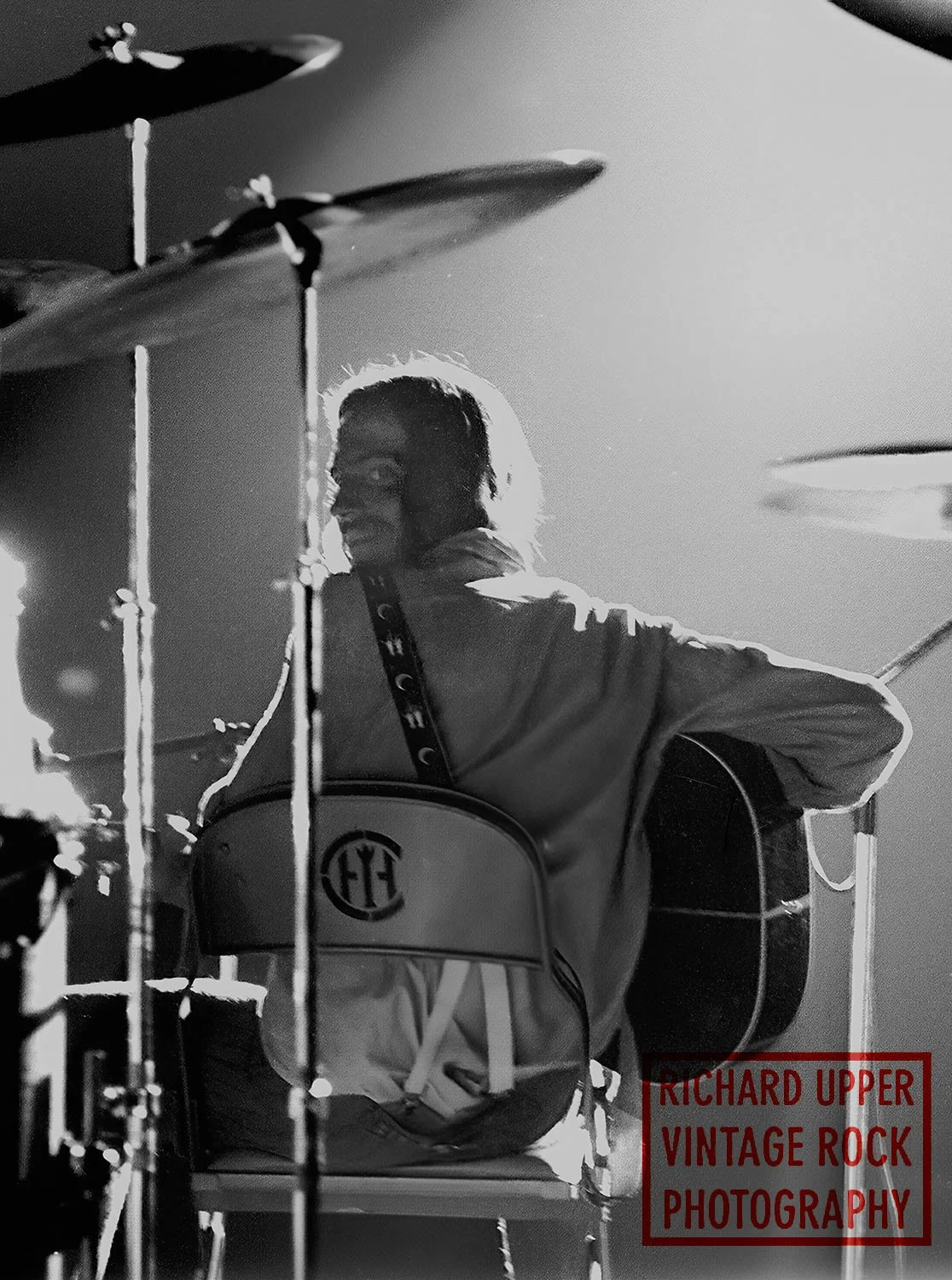 A person sitting on a chair, playing an acoustic guitar, with a drum set in the foreground, black and white photo.
