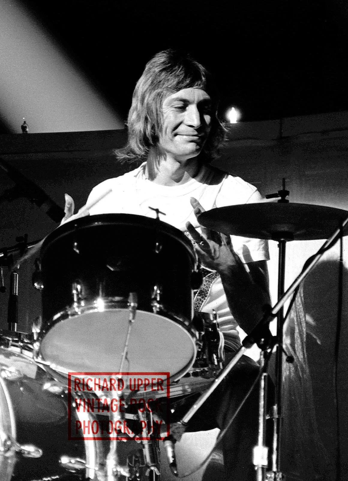 A black and white photo of a person with shoulder-length hair playing drums, smiling and looking down at the drum set.