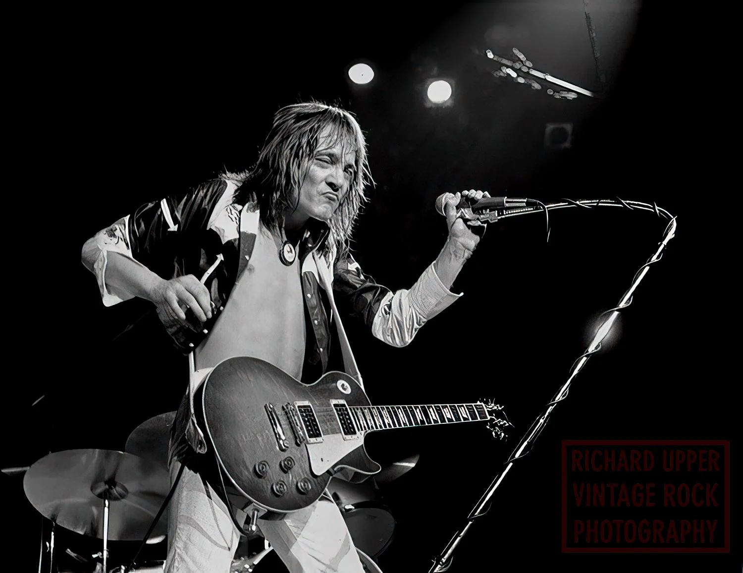 Black and white photo of a long-haired male guitarist on stage, holding a microphone in one hand and a guitar with the other, with a focused expression.