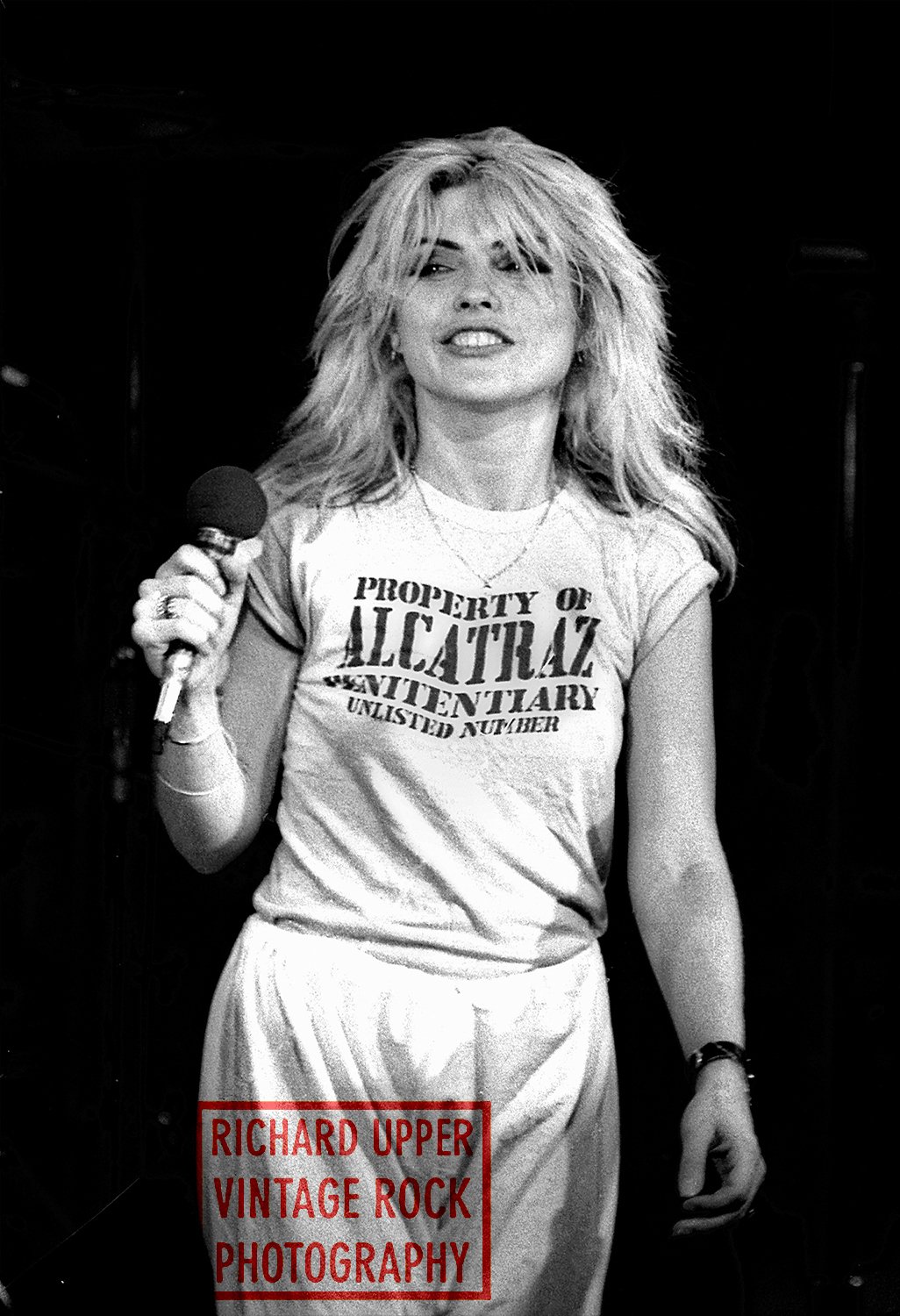 Black and white photograph of a woman with voluminous, messy blonde hair, holding a microphone in her right hand, wearing a t-shirt with text, and standing against a dark background, suggesting a vintage rock performance or candid moment.
