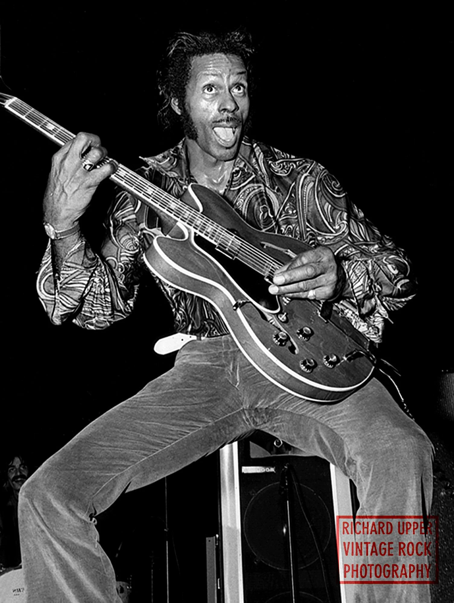 Black and white photo of a man with medium-length hair and sideburns playing an electric guitar, wearing a patterned shirt and bell-bottom jeans, with a joyful expression and mouth open.