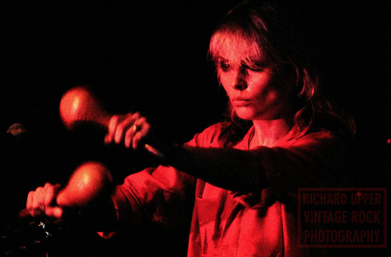A woman with blonde hair and focused expression playing an electric guitar in a dark setting, lit by red lighting.