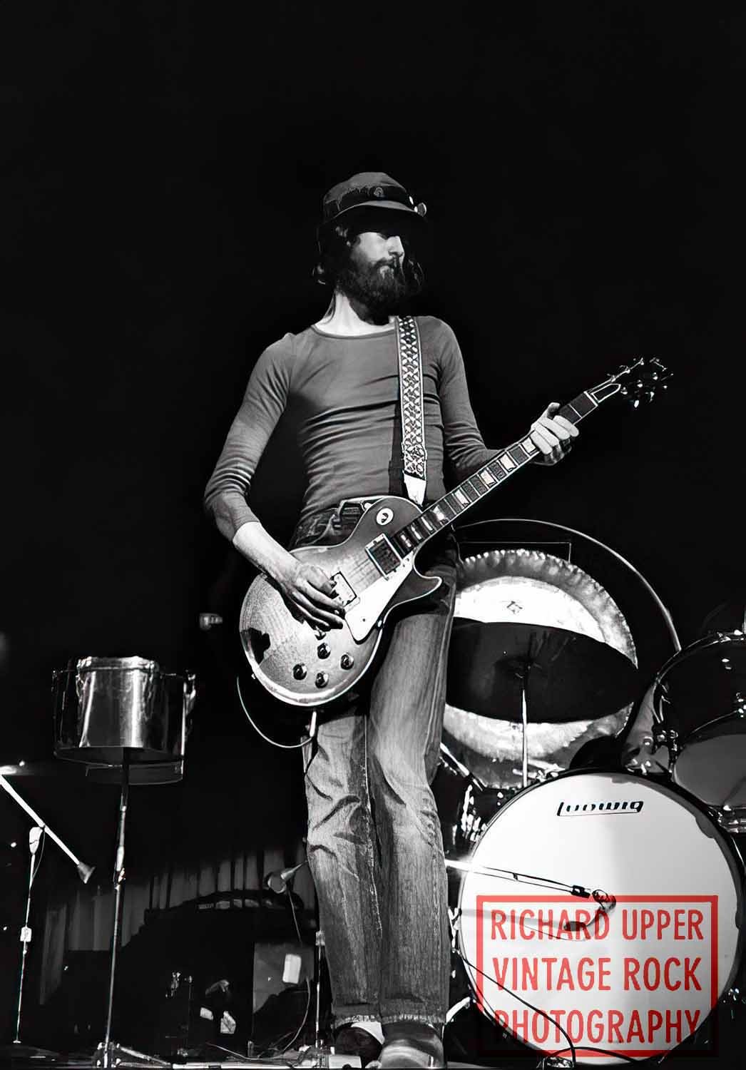 A man with a beard, wearing a hat and long-sleeved shirt, playing an electric guitar on stage with musical drums behind him. The setting appears to be a concert or live music performance.