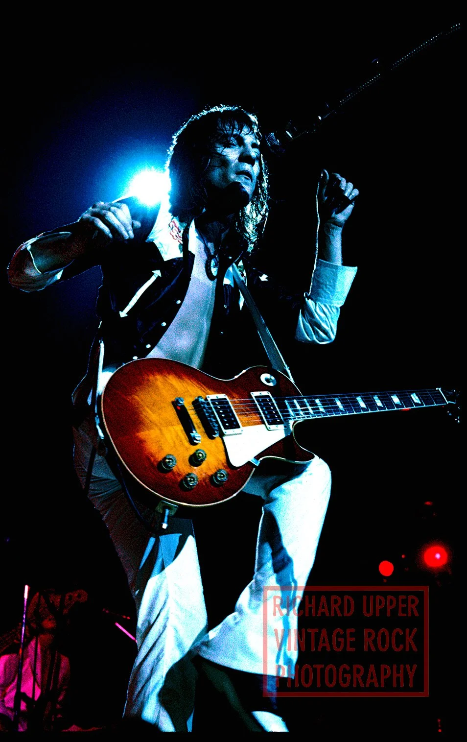 A male guitarist performing on stage with a sunburst electric guitar, dark background, bright stage lights, and clenched fist.