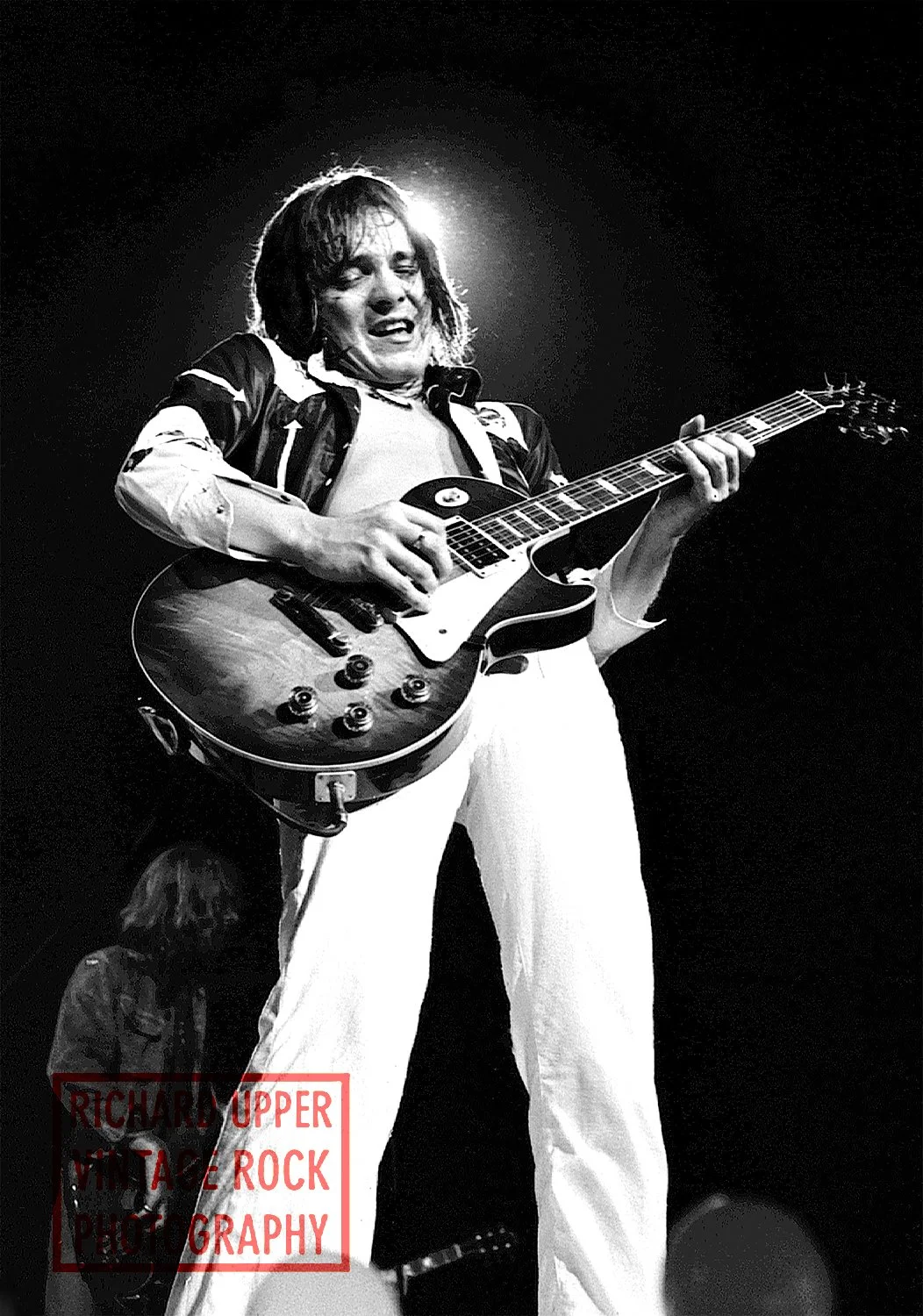 Black and white photo of a musician playing an electric guitar on stage, with intense facial expression, backlit with a halo effect around his head, and another person partially visible in the background.