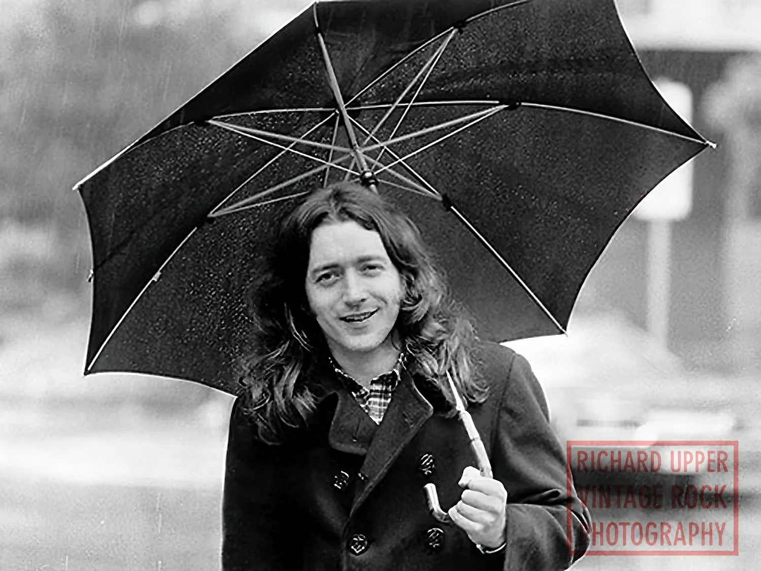 A person with long hair, smiling, holds an umbrella in the rain.