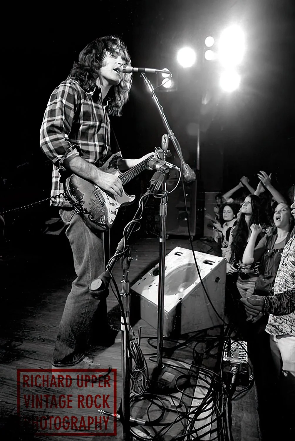 A black and white photo of a vintage rock concert featuring a male musician with long, curly hair, wearing a plaid shirt, playing an electric guitar and singing into a microphone on stage. The band is performing with an enthusiastic audience visible 