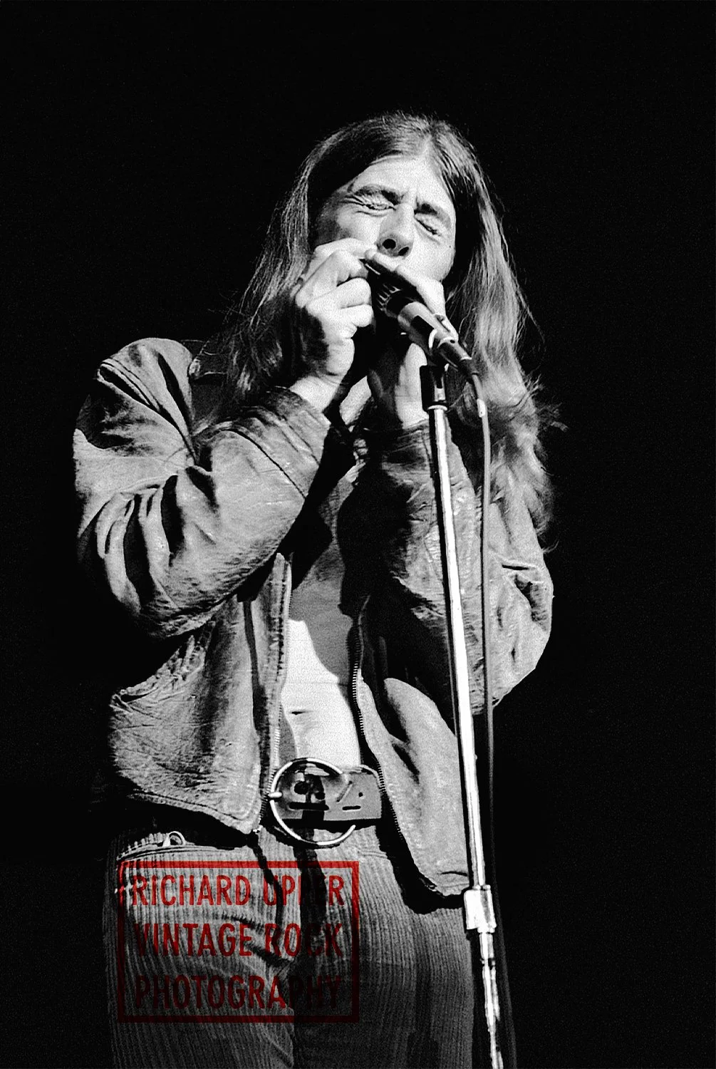 Black and white photo of a male singer with long hair, wearing a leather jacket, singing into a microphone on stage.