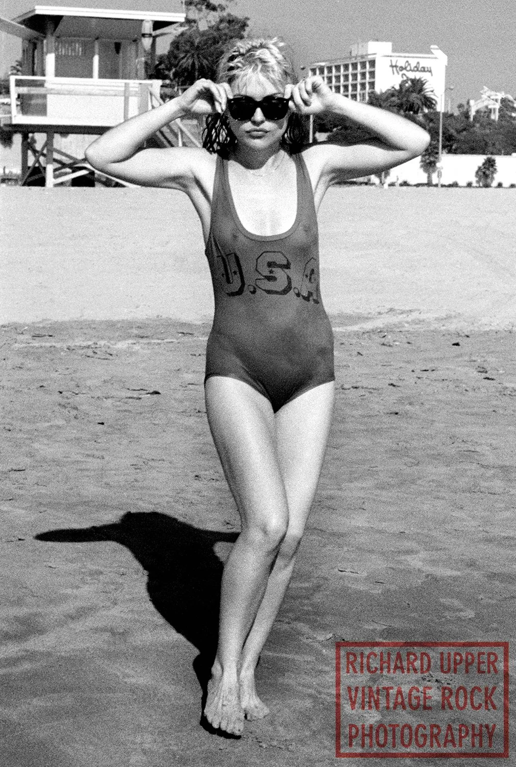 A woman wearing sunglasses and a vintage one-piece swimsuit with the letters 'U.S.' printed on it, standing barefoot on the beach and adjusting her sunglasses, with lifeguard towers and a sign in the background.