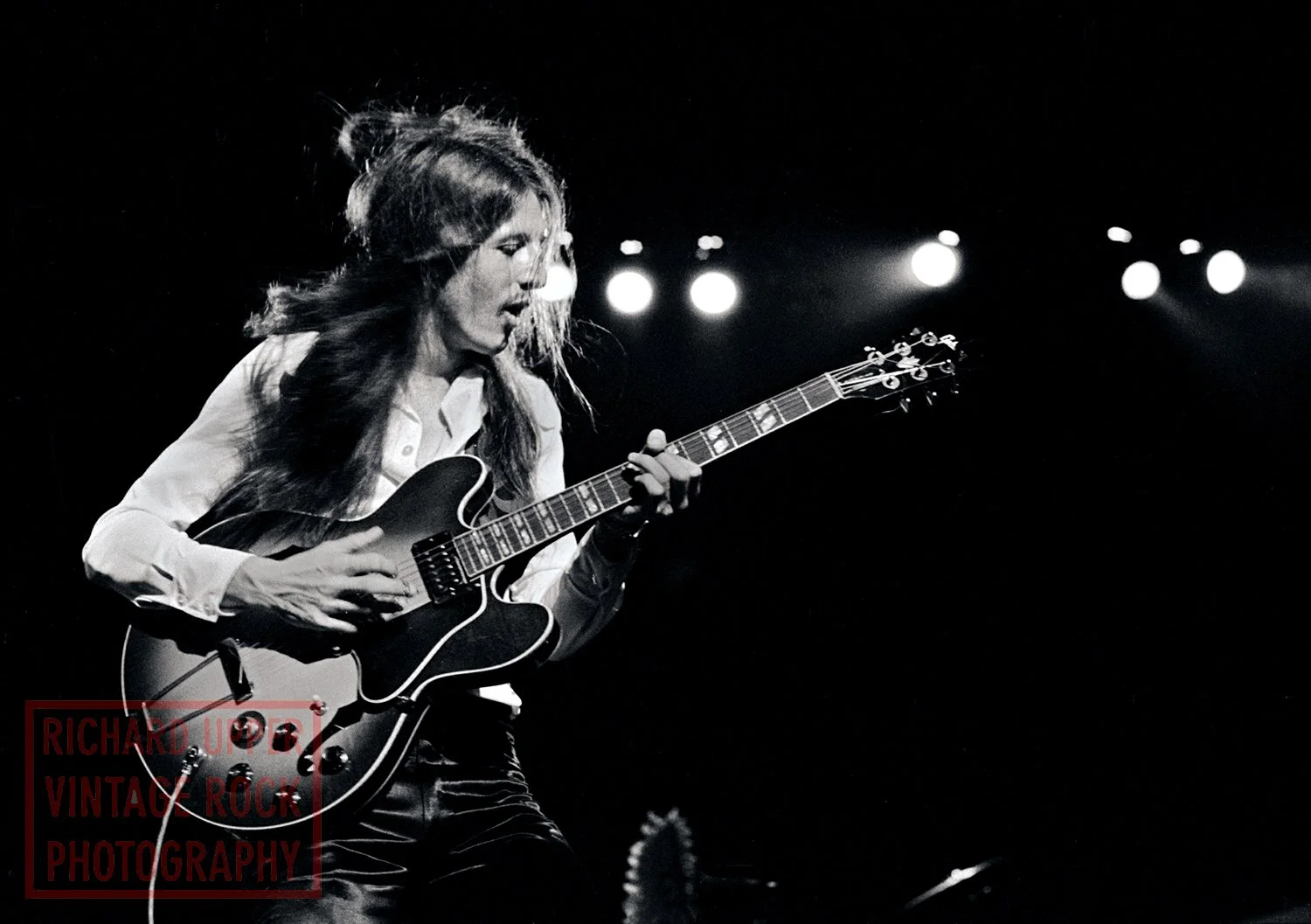 A woman with long hair playing an electric guitar on stage, with stage lights behind her, captured in black and white.