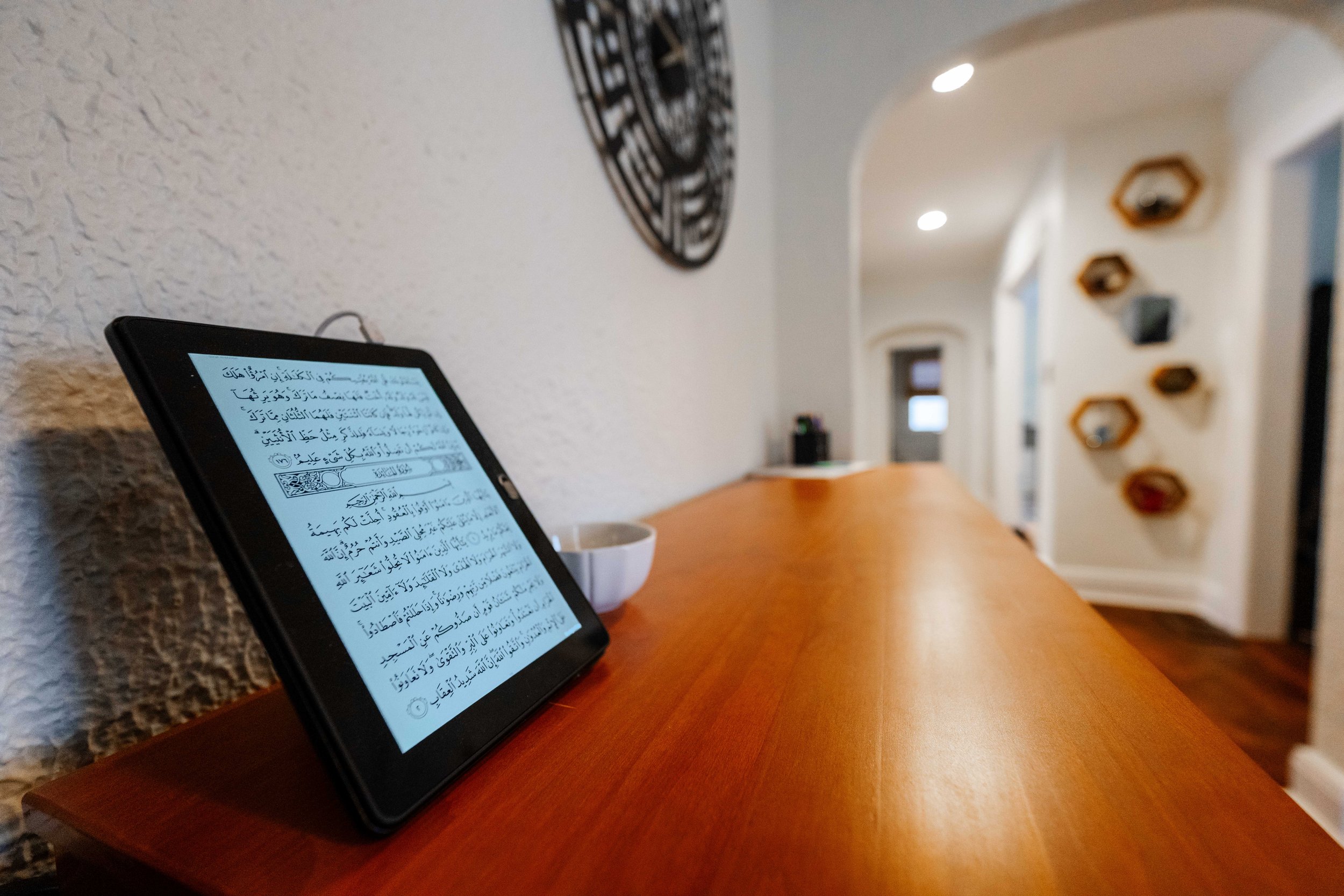 Digital tablet on a wooden counter displaying Arabic text from the Qur'an, with a white bowl beside it, in a cozy room with textured white walls, decorative wall art, and shelves.