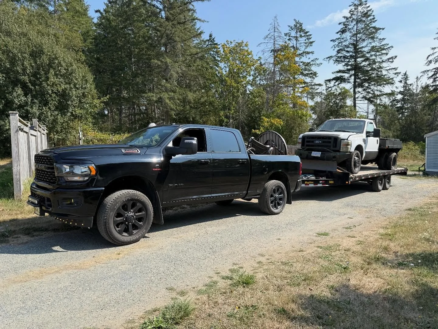 A black pickup truck towing a flatbed trailer with a white truck on it, parked on a gravel driveway surrounded by trees and a wooden fence.