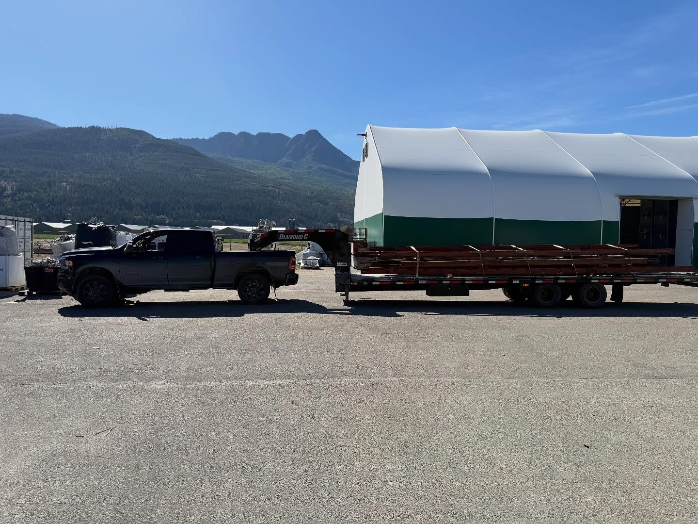 A black pickup truck towing a flatbed trailer with benches in front of a large white and green tent structure, with mountains in the background and a clear blue sky.