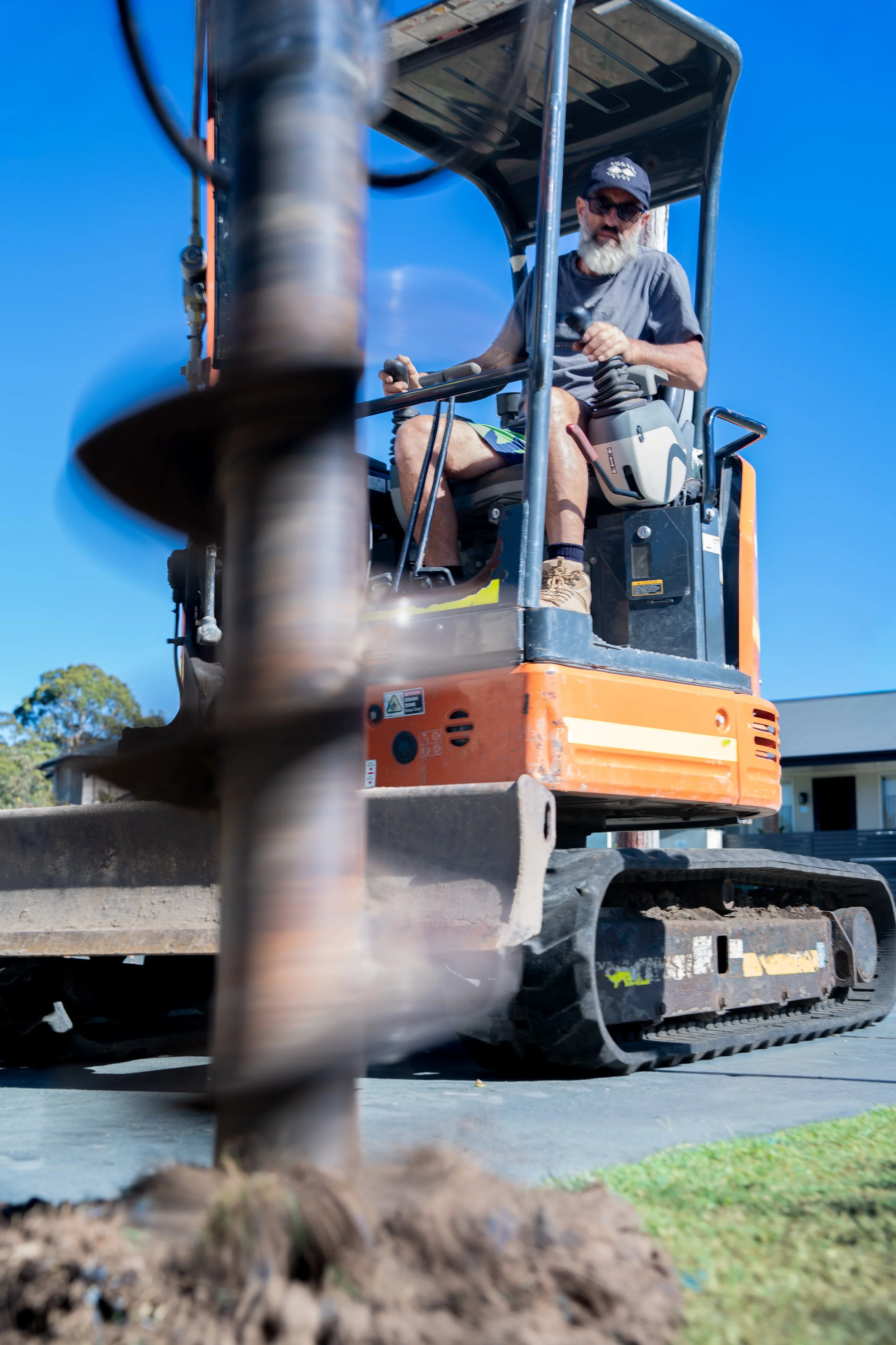 A man operating an orange compact excavator with a drilling attachment outdoors on a sunny day.