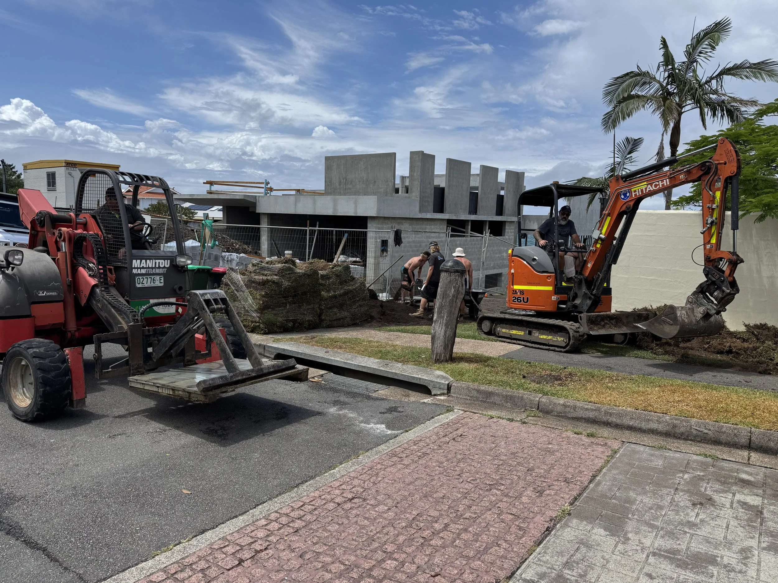 Construction workers using heavy machinery to dig and level the ground outside a building under construction, with a blue sky and scattered clouds above.