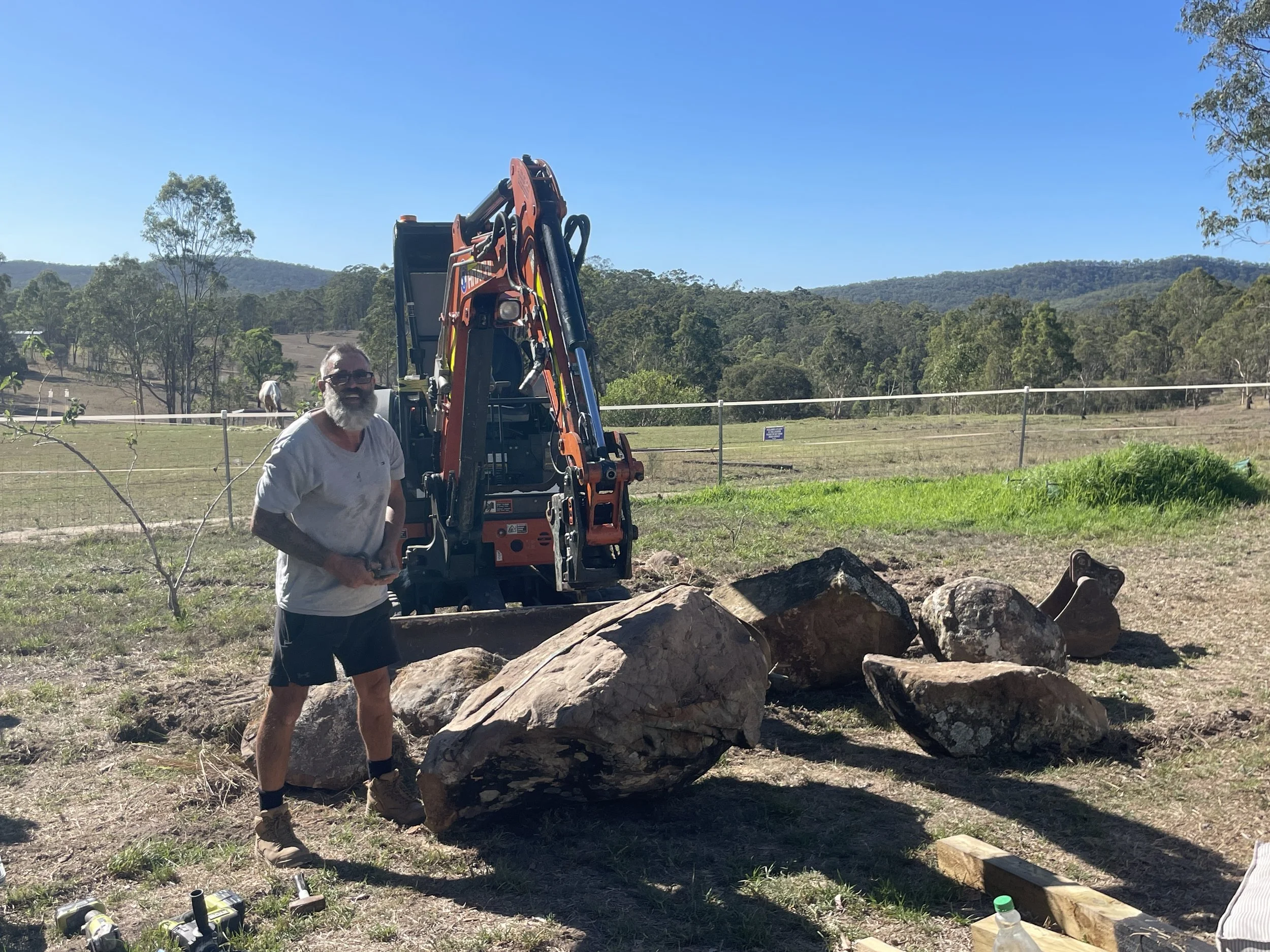 A man with a beard and glasses standing outdoors near a small excavator and large rocks. The background shows a rural landscape with trees and hills under a clear blue sky.