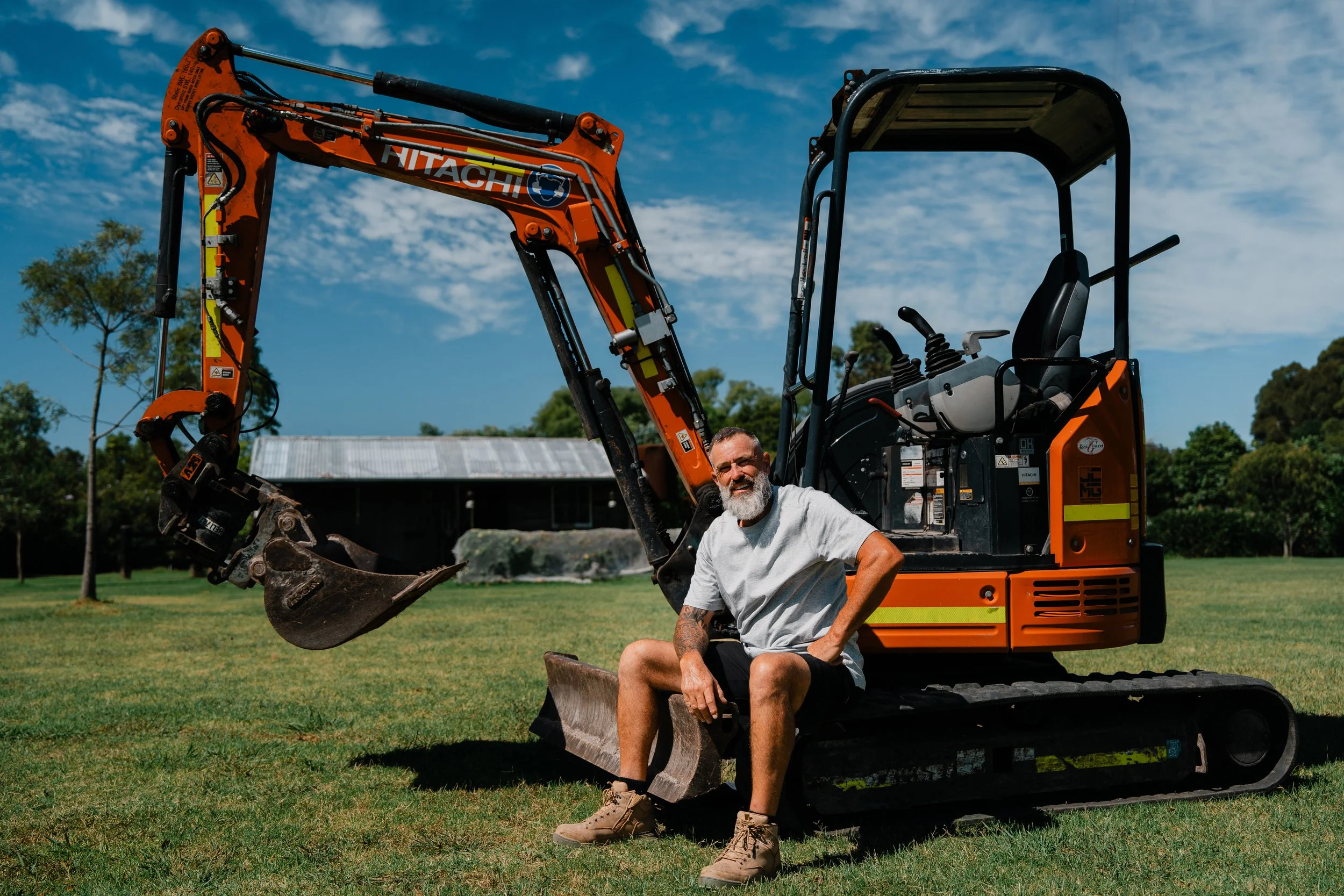 A man with a beard and tattoos sitting on a small excavator on a grassy field under a partly cloudy sky.