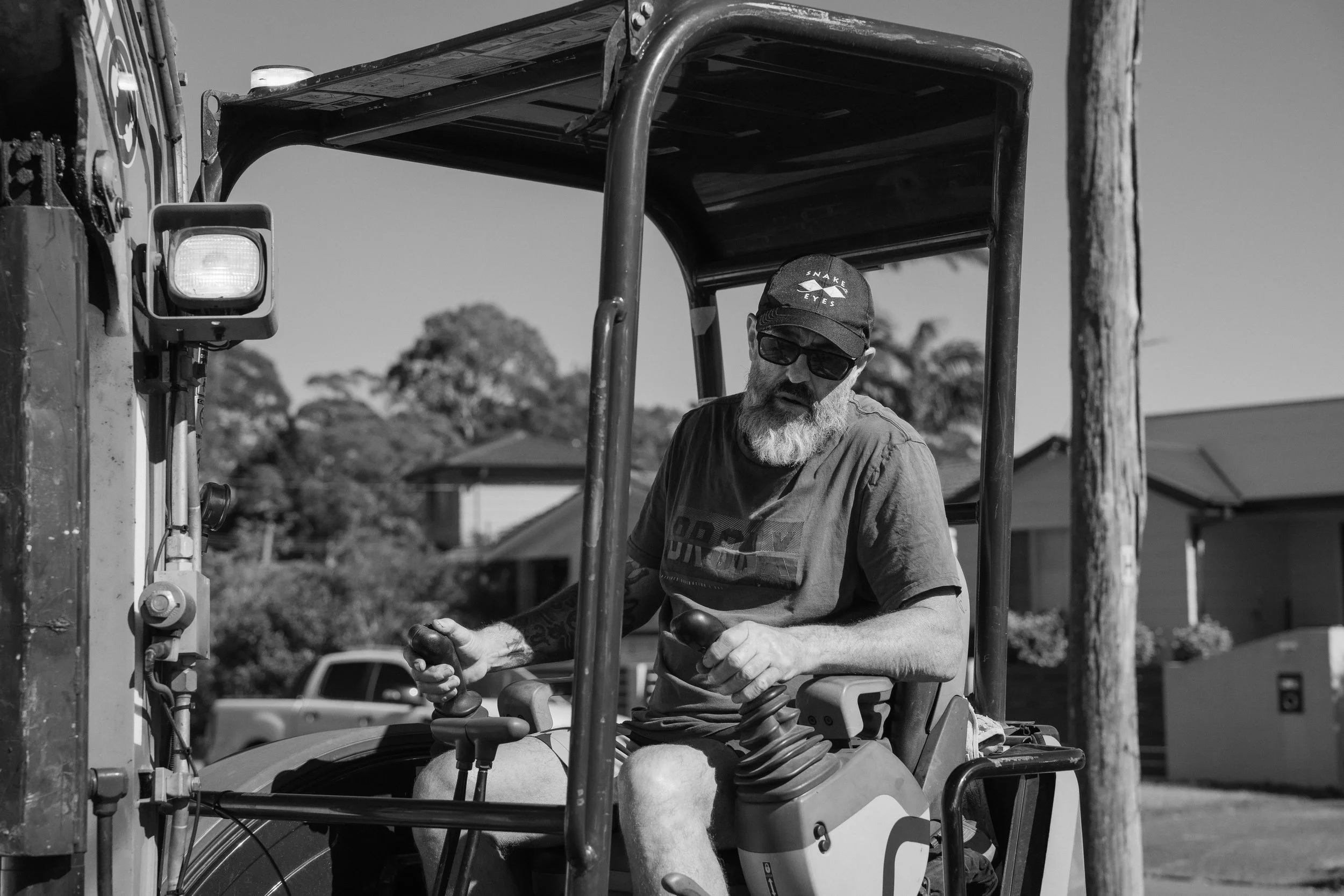 A man with a beard, sunglasses, and a cap operating a small construction vehicle on a residential street.