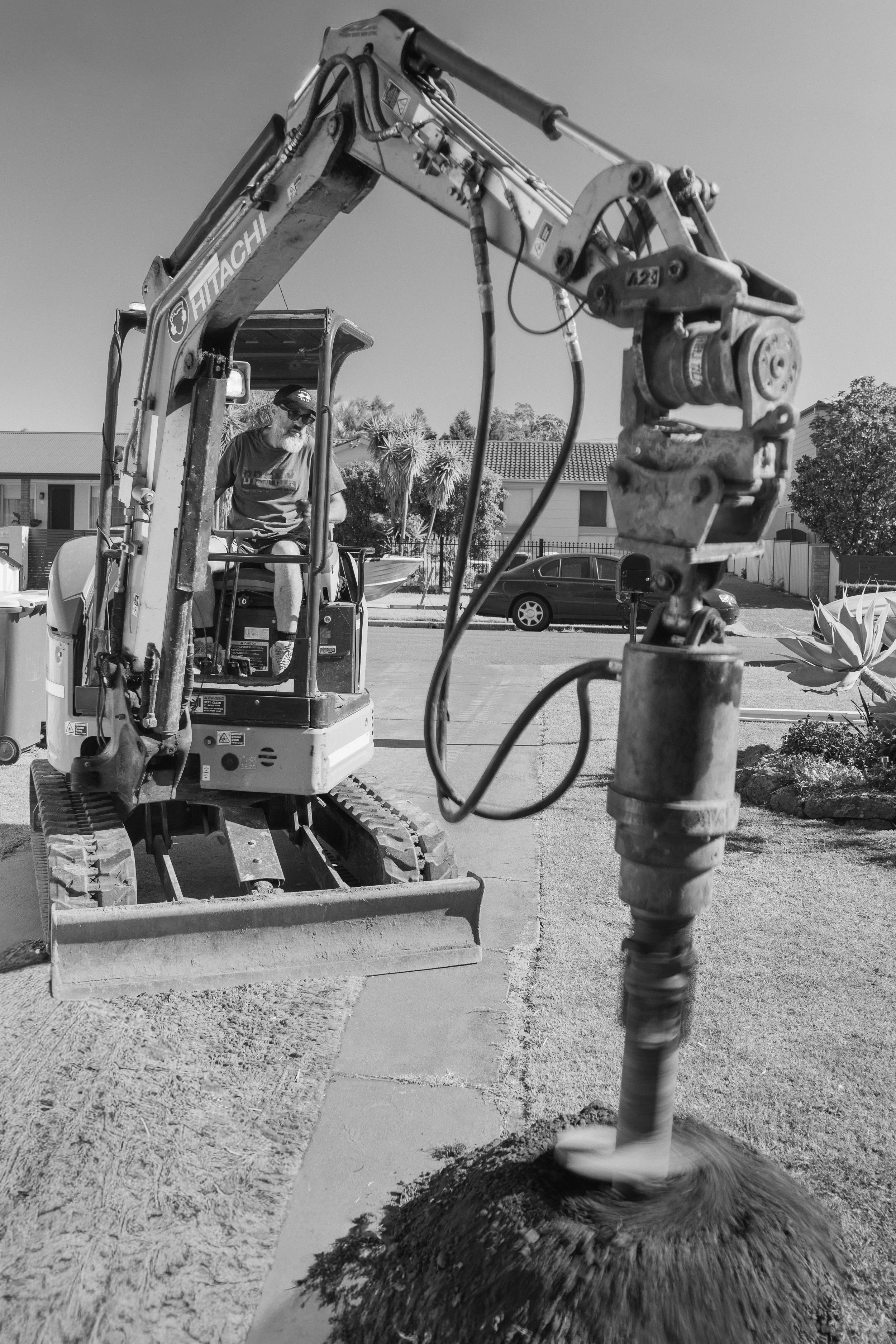 Black and white photo of a man operating a small excavator on a residential street, with houses and parked cars in the background.
