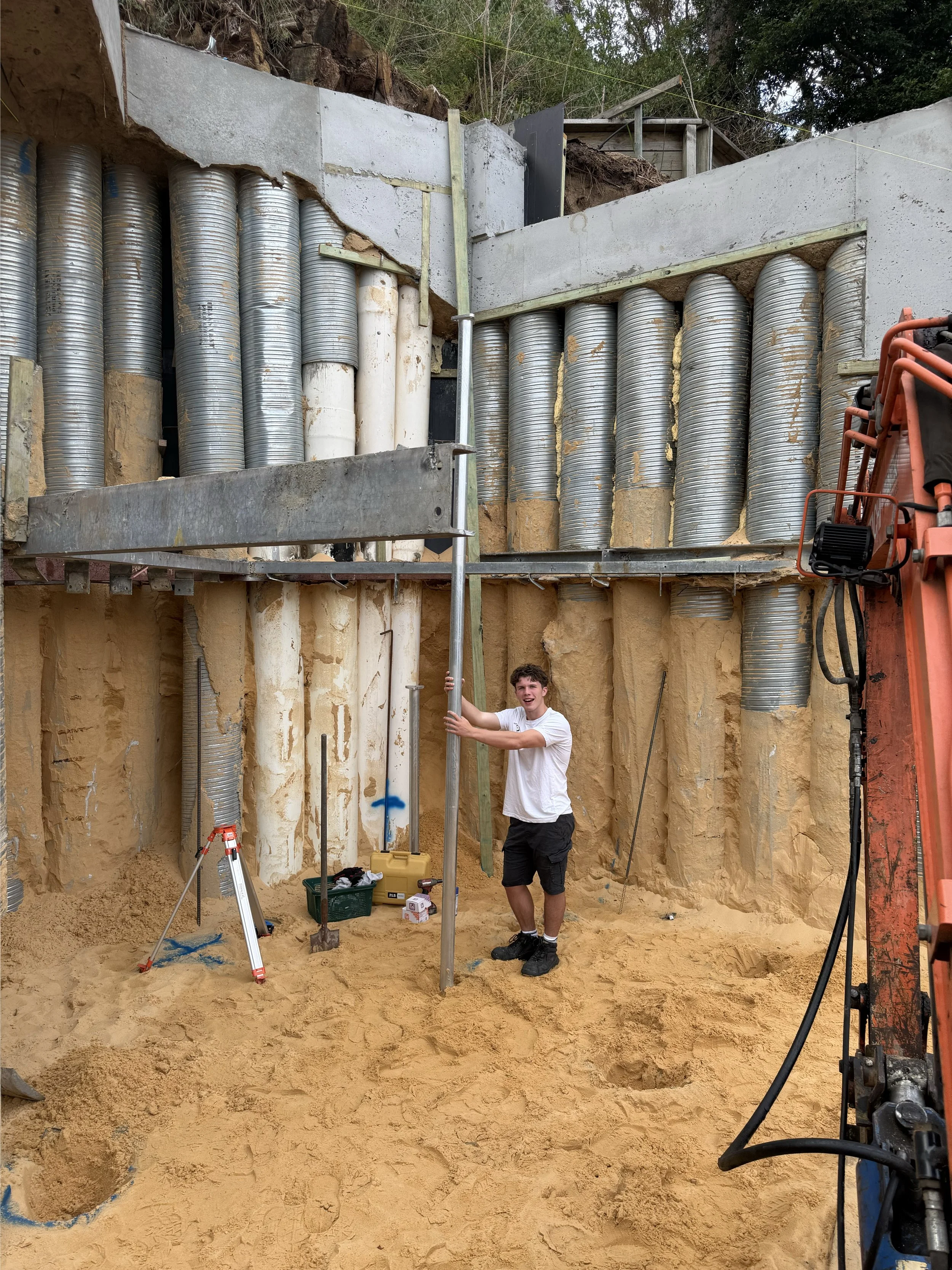 A construction worker smiling and holding a pole at an underground construction site with large ventilation pipes and sand walls.