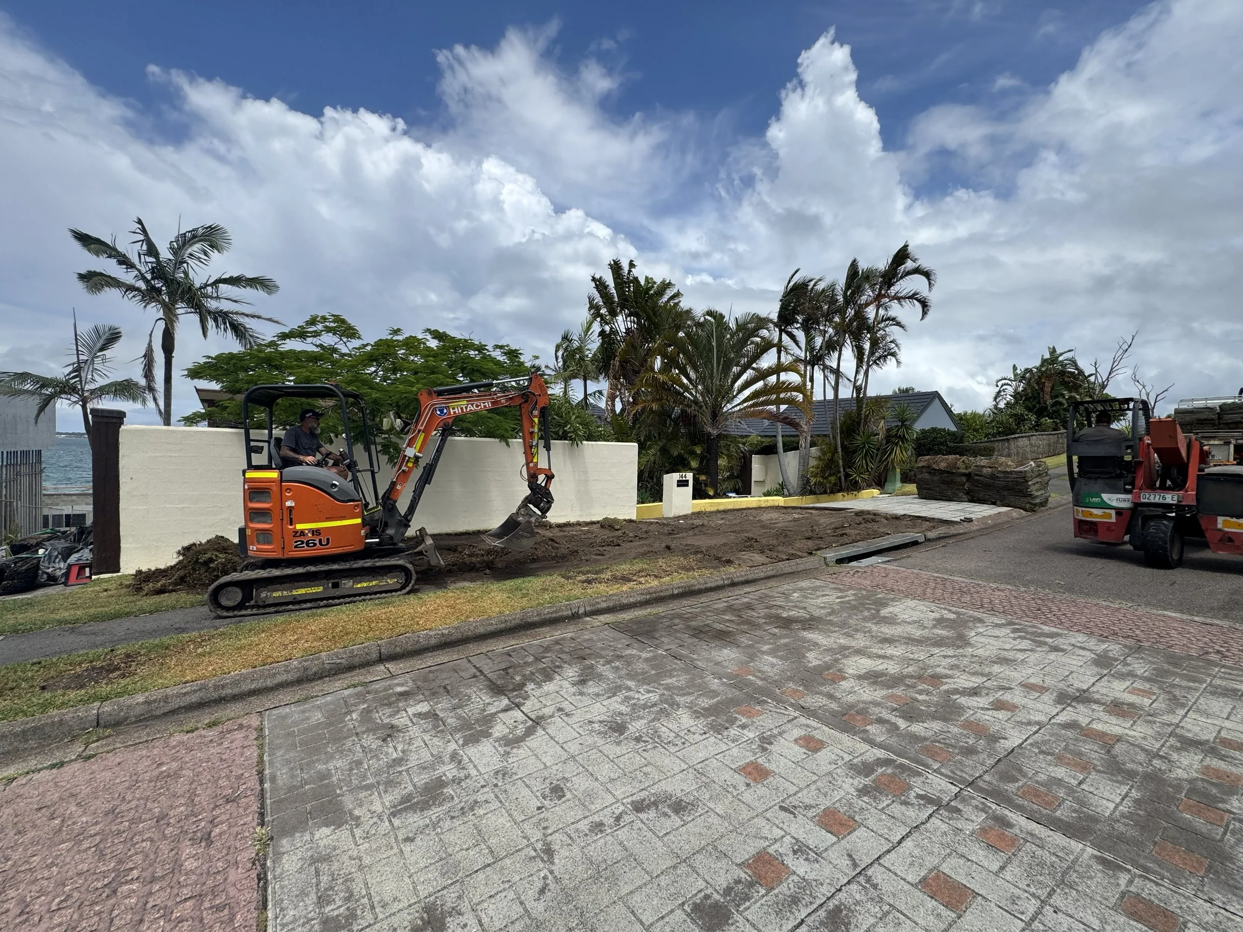 Construction workers operating a mini excavator on a residential street, with trees, a white wall, and houses in the background under a partly cloudy sky.