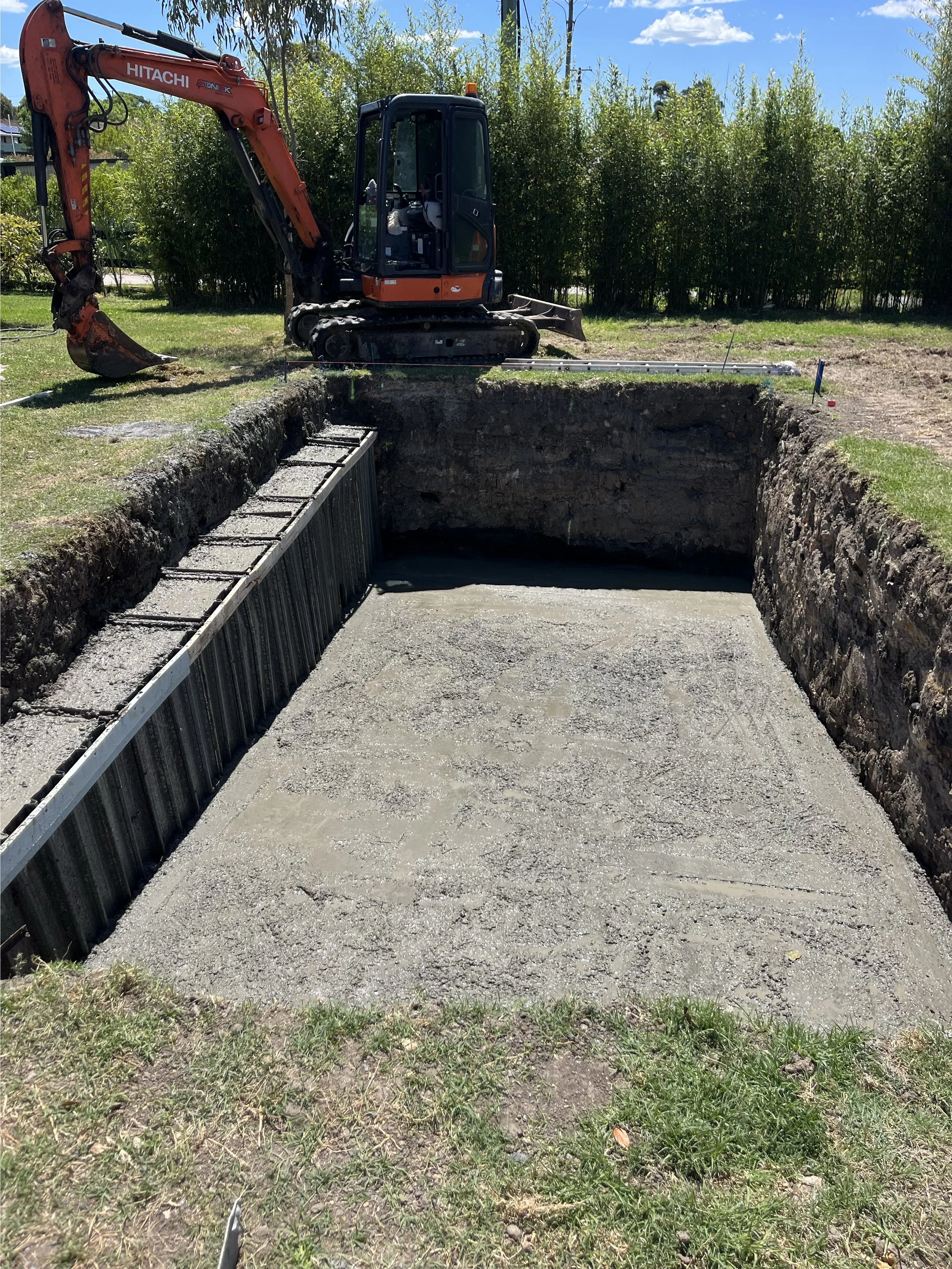Construction site with an excavator digging a large rectangular hole in the ground, with the floor leveled and reinforced with metal signs on the sides. The background features trees and a partly cloudy sky.