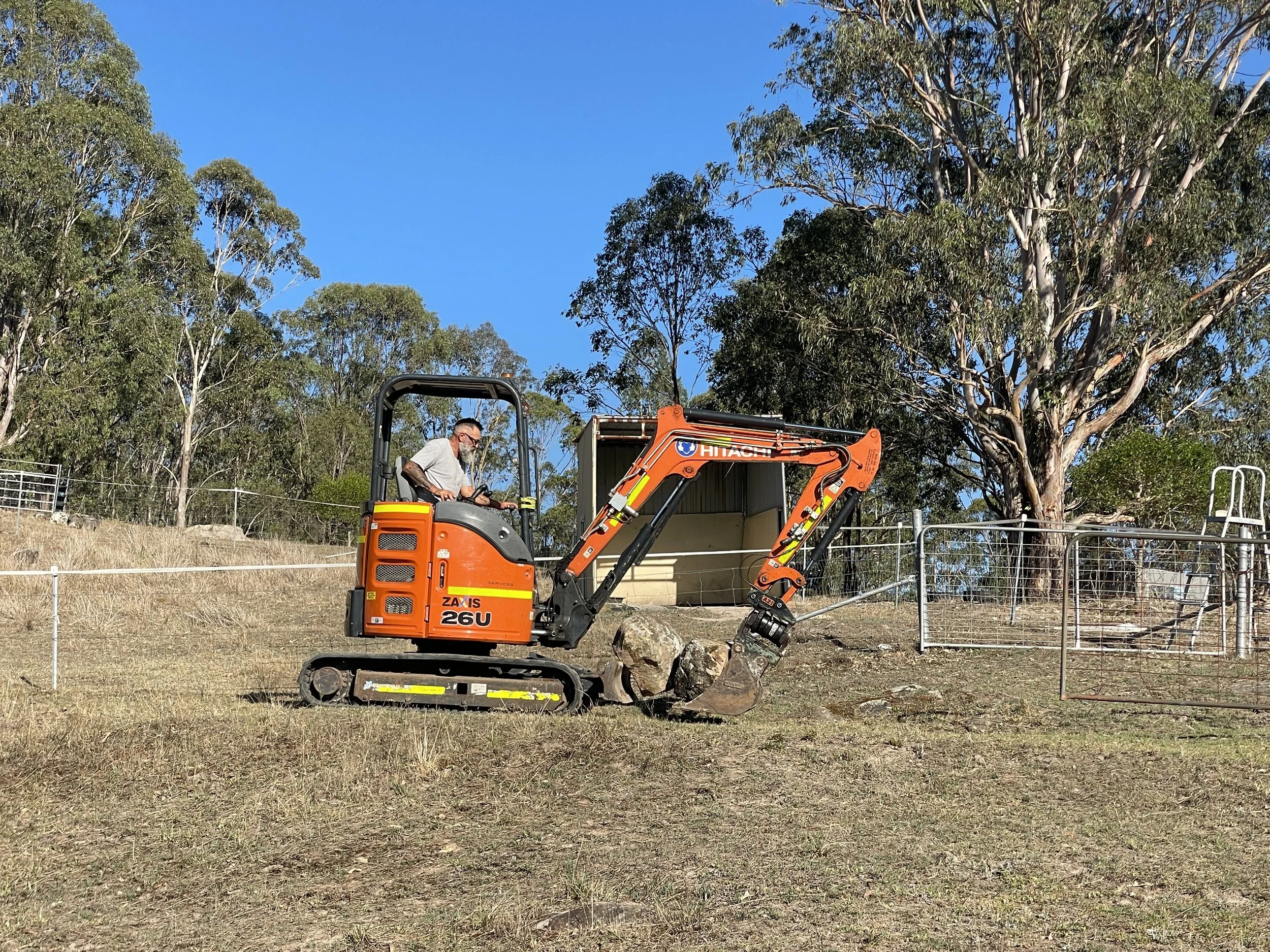 A person operating an orange Hitachi mini excavator on a grassy field with trees and a small shed in the background, under a clear blue sky.