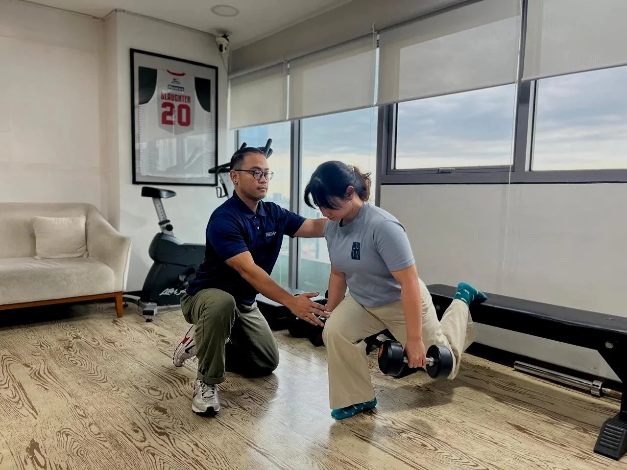 A female patient doing a split on a workout bench while holding a dumbbell in each hand, being assisted by a male trainer in a fitness room with large windows, a framed sports jersey on the wall, and exercise equipment.