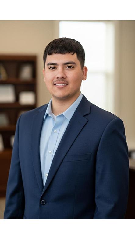 A young man in a navy suit and light blue shirt standing in an office with large windows and bookshelf