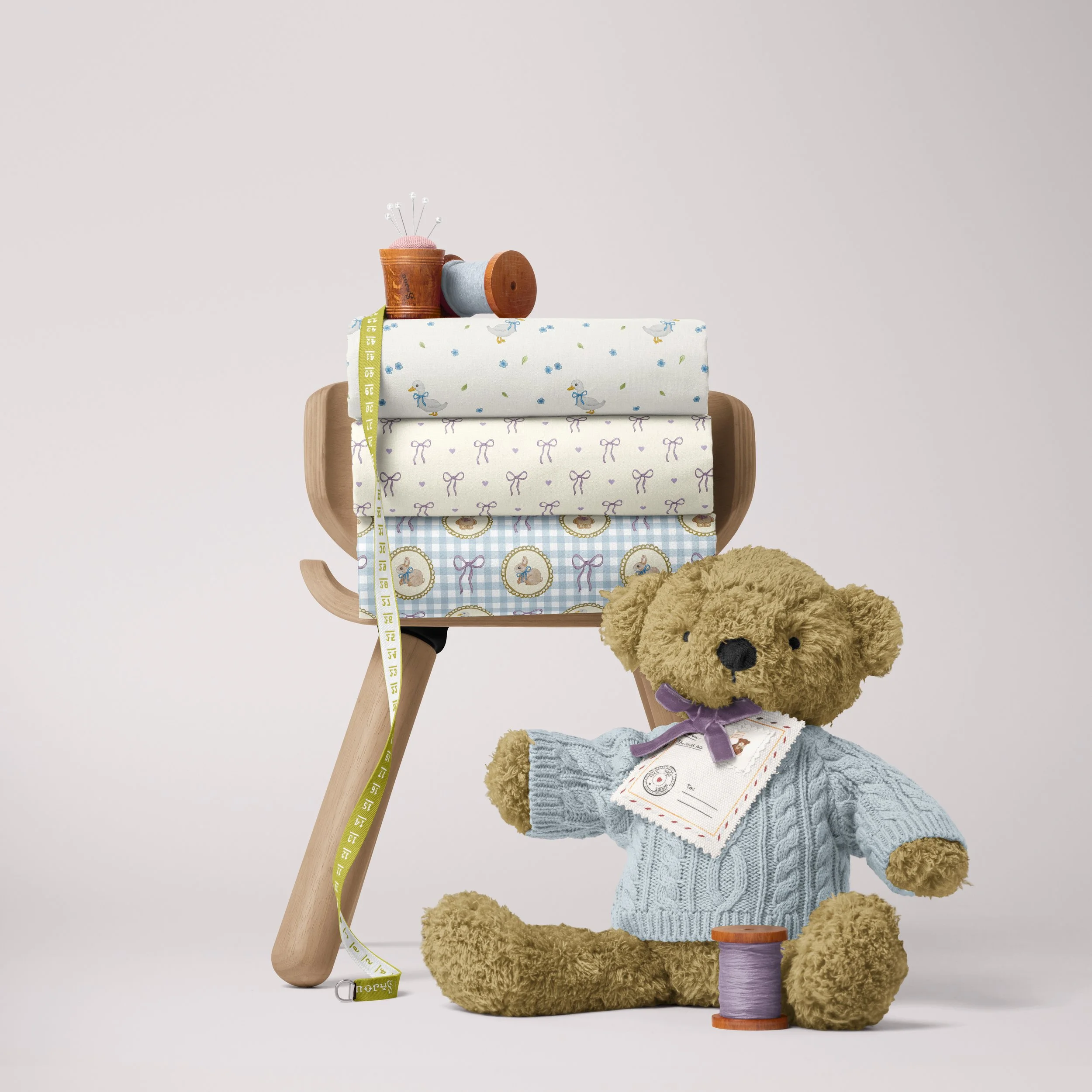 A teddy bear wearing a sweater and purple ribbon sitting next to sewing supplies including fabric, a measuring tape, and thread, with sewing tools on a wooden table.