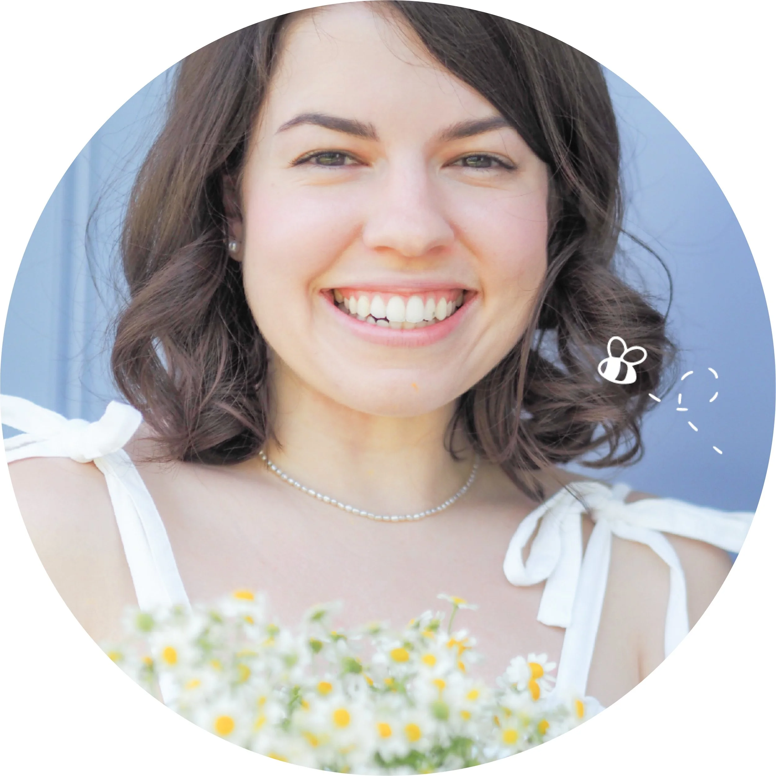 A woman with short brown hair, smiling, wearing a white dress with shoulder ties, a pearl necklace, and small earrings, holding a bouquet of small white and yellow flowers, outdoors with a blue background including a drawing of a bee.