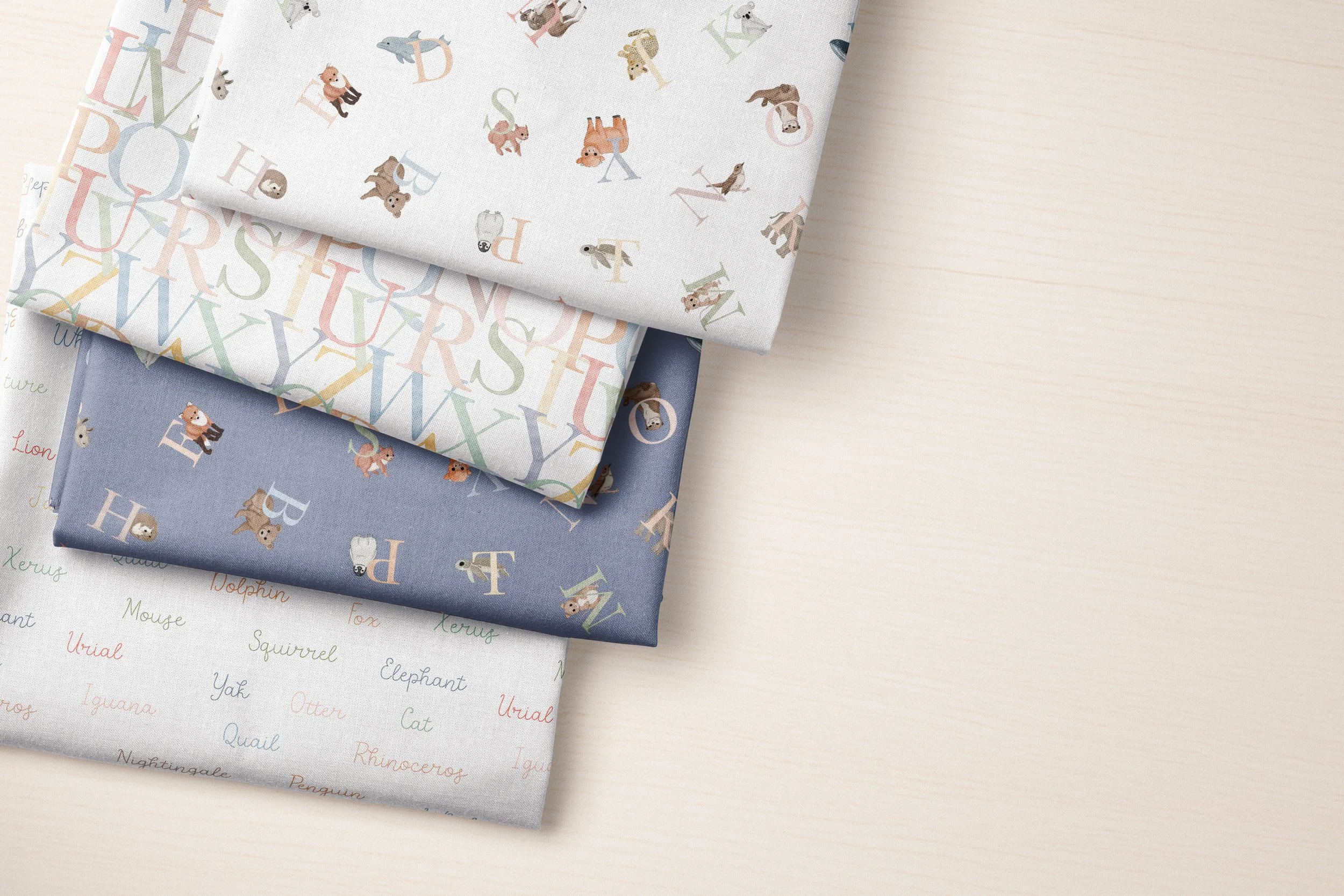 Stack of five folded patterned fabrics featuring animals, letters, and alphabet words on a light-colored surface.