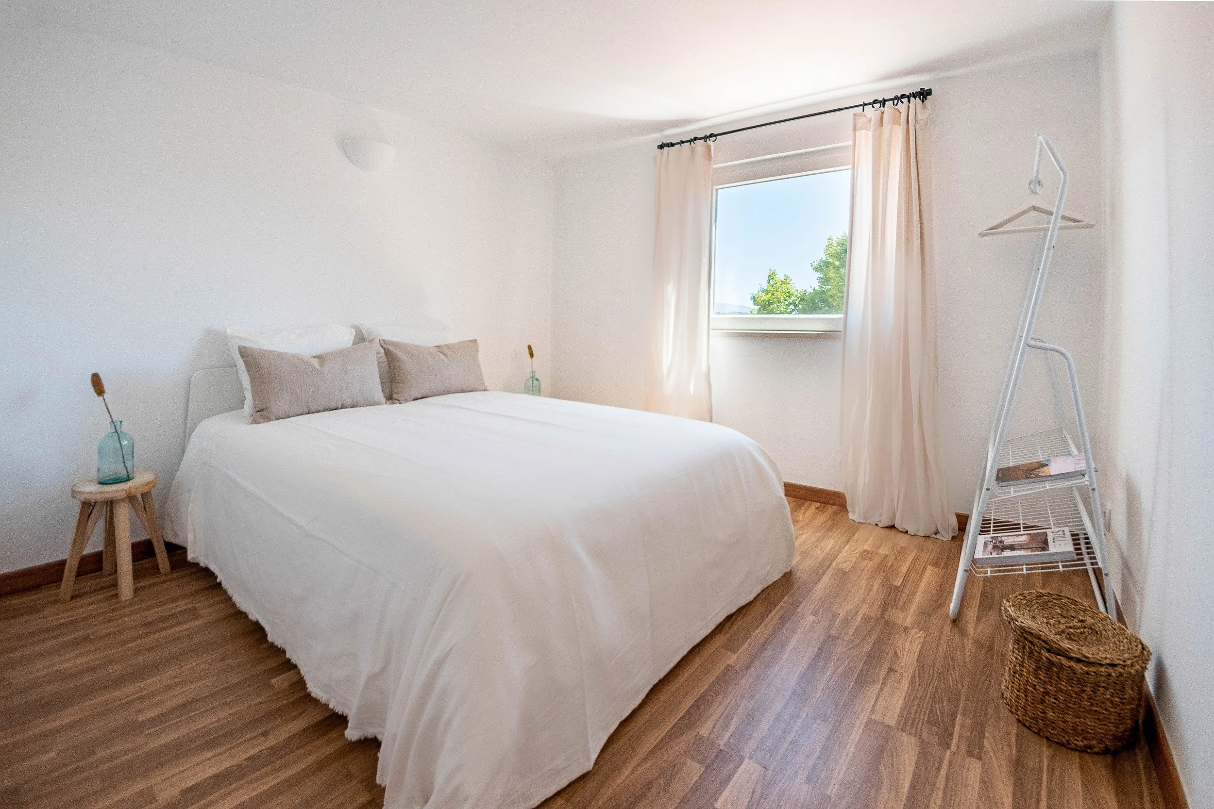 A minimalist bedroom with a white bed, two beige pillows, wooden side tables with glass vases, a window with cream curtains, a foldable clothing rack, and a woven basket, with wooden flooring and white walls.