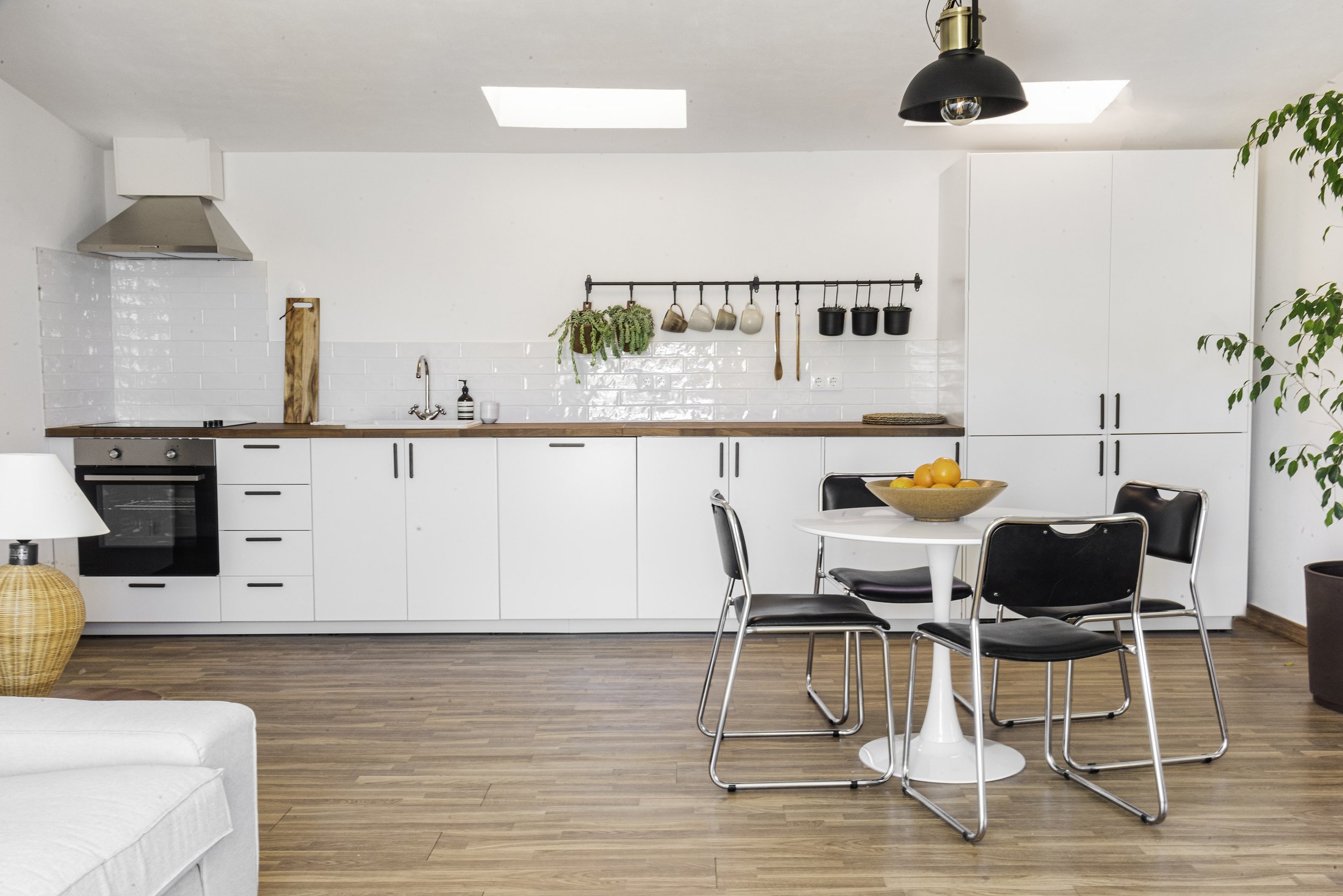 Modern kitchen with white cabinetry, black and gold appliances, wooden countertop, and a small round dining table with four black chairs, a bowl of lemons, and a hanging rail with pots, pans, and plants.