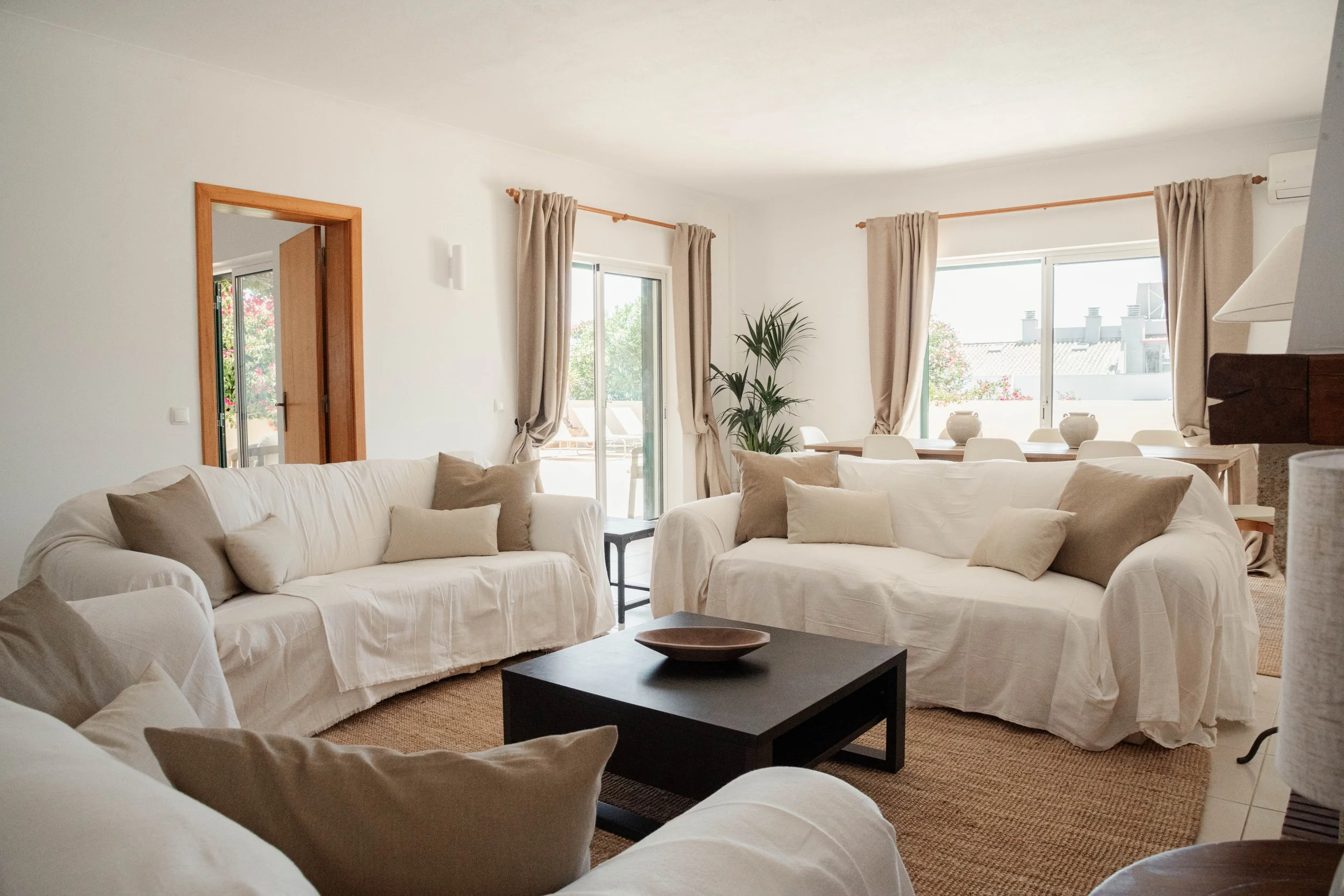 A minimalist bedroom with a white bed, two beige pillows, wooden side tables with glass vases, a window with cream curtains, a foldable clothing rack, and a woven basket, with wooden flooring and white walls.