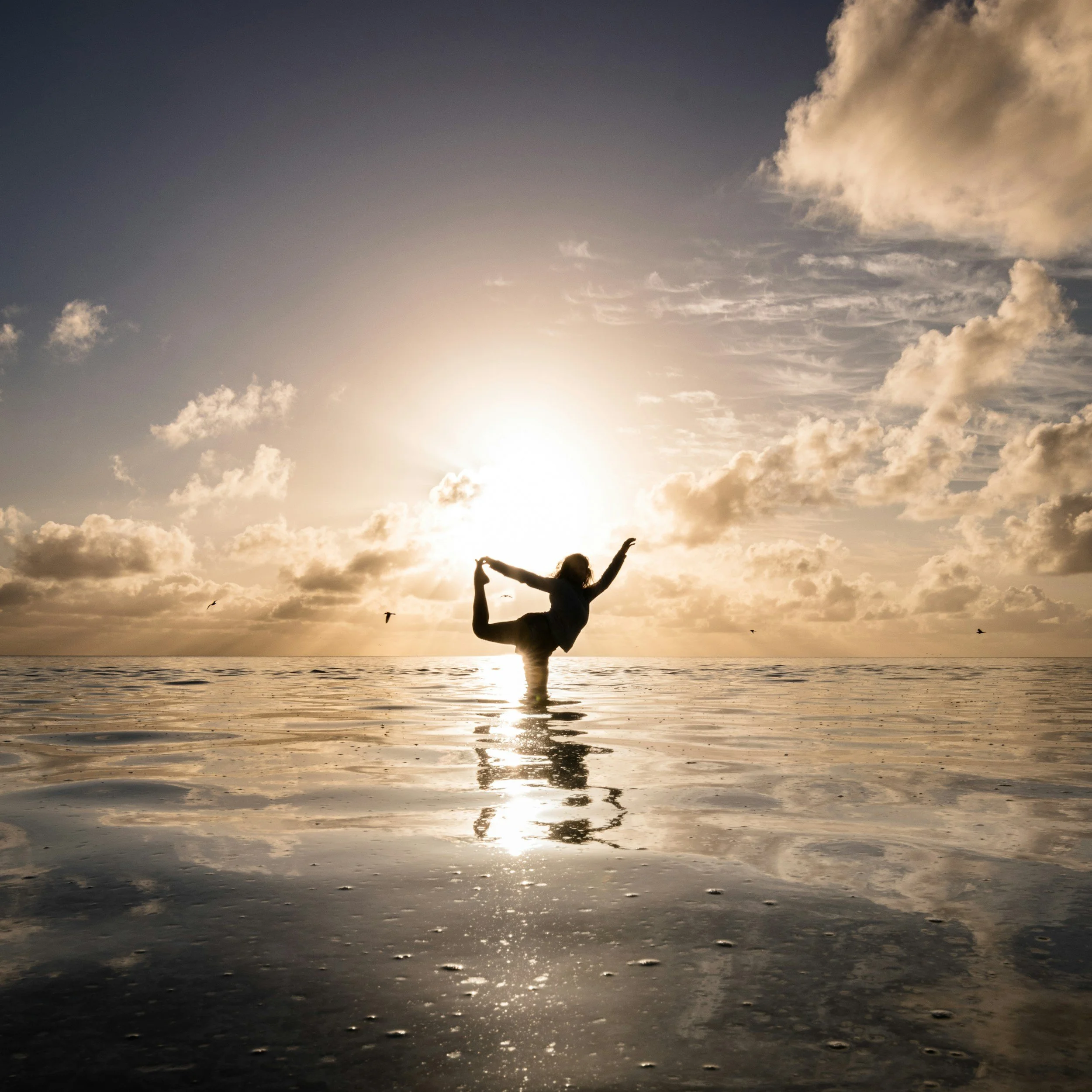 Person performing a yoga pose with arms raised at the beach during sunset, with clouds and seagulls in the sky.