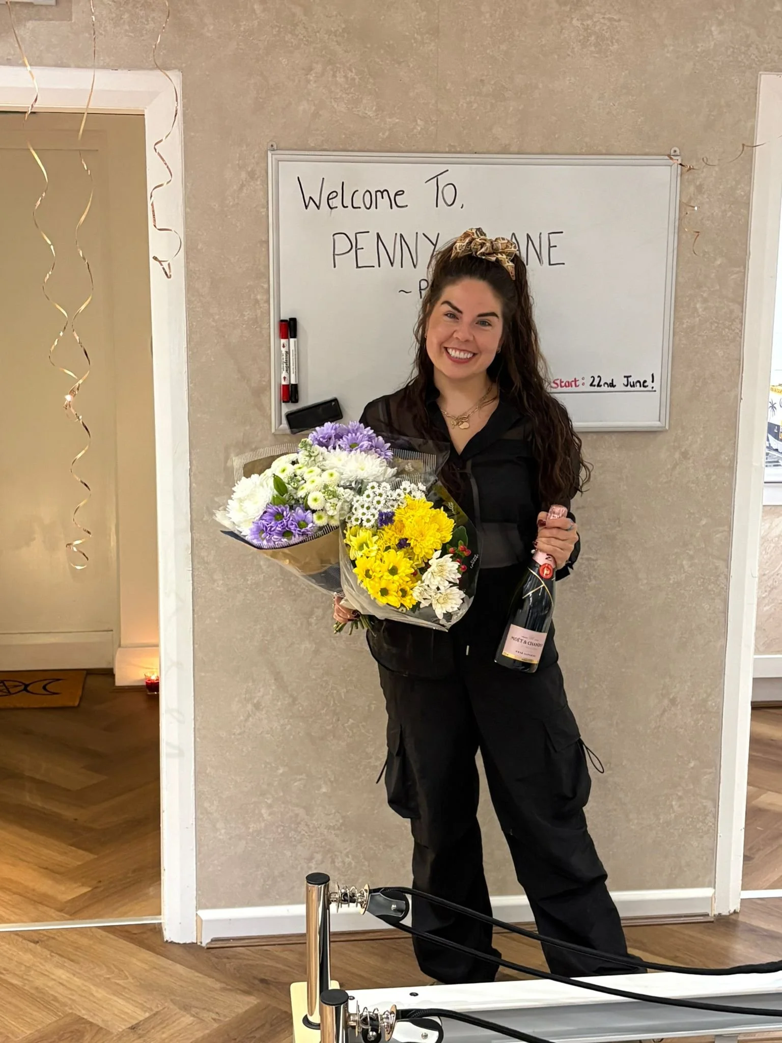 A woman standing indoors, smiling, holding a large bouquet of flowers and a bottle of champagne. Behind her is a whiteboard with the words 'Welcome To Penny Lane' and an event date. Decorations are visible around the doorway.